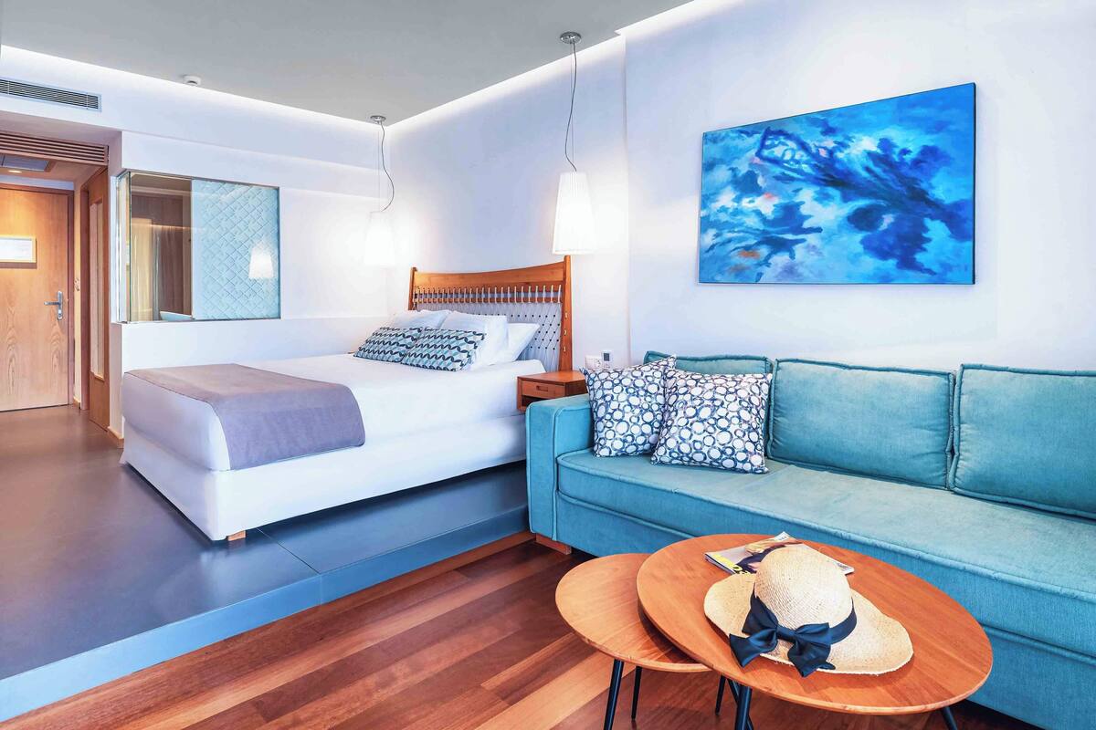Una habitación de hotel moderna con una cama, un sofá azul, una pequeña mesa con un sombrero y un cuadro en la pared.