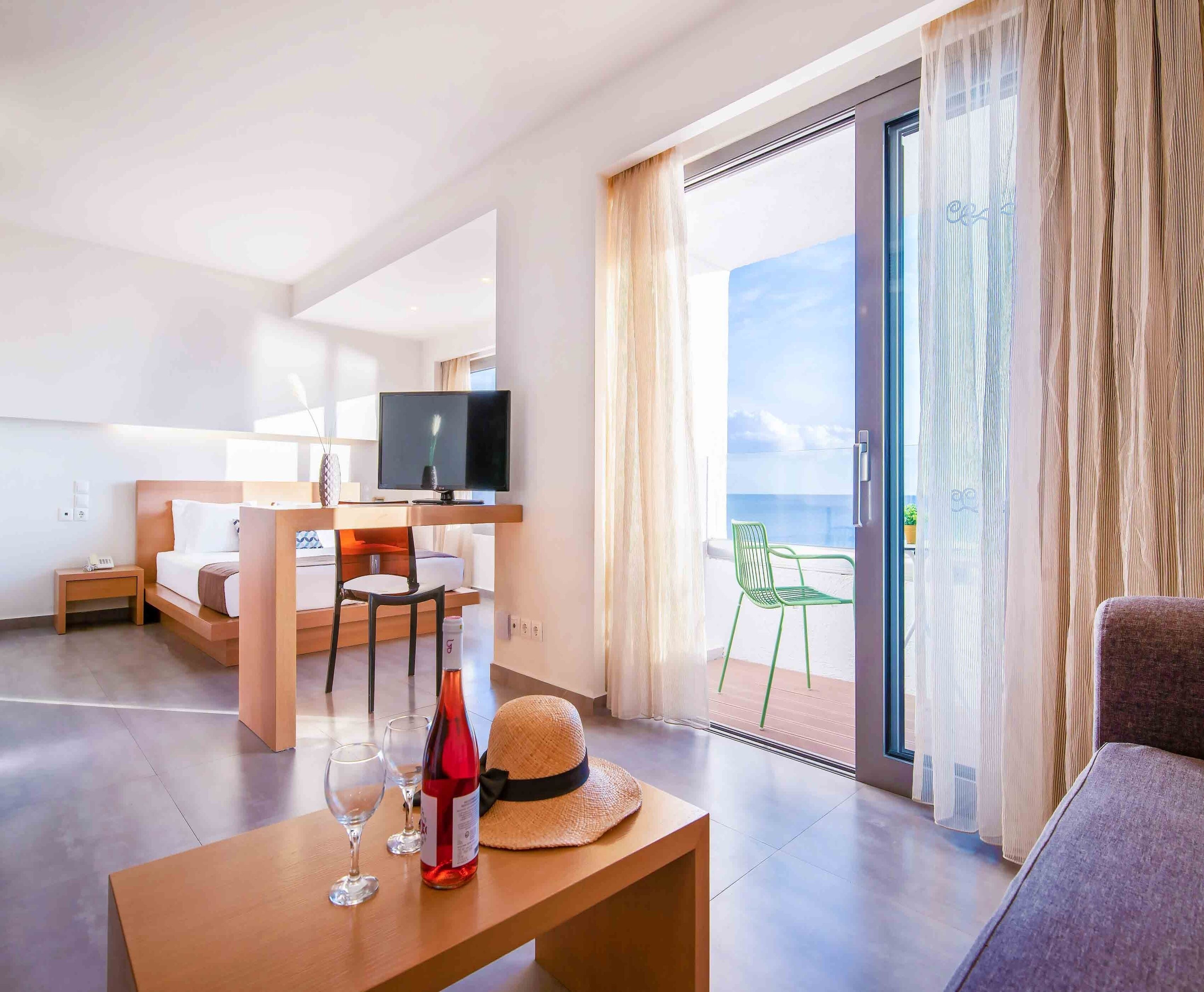 A modern hotel room with a balcony, a television, a dining area, and a bottle of wine.