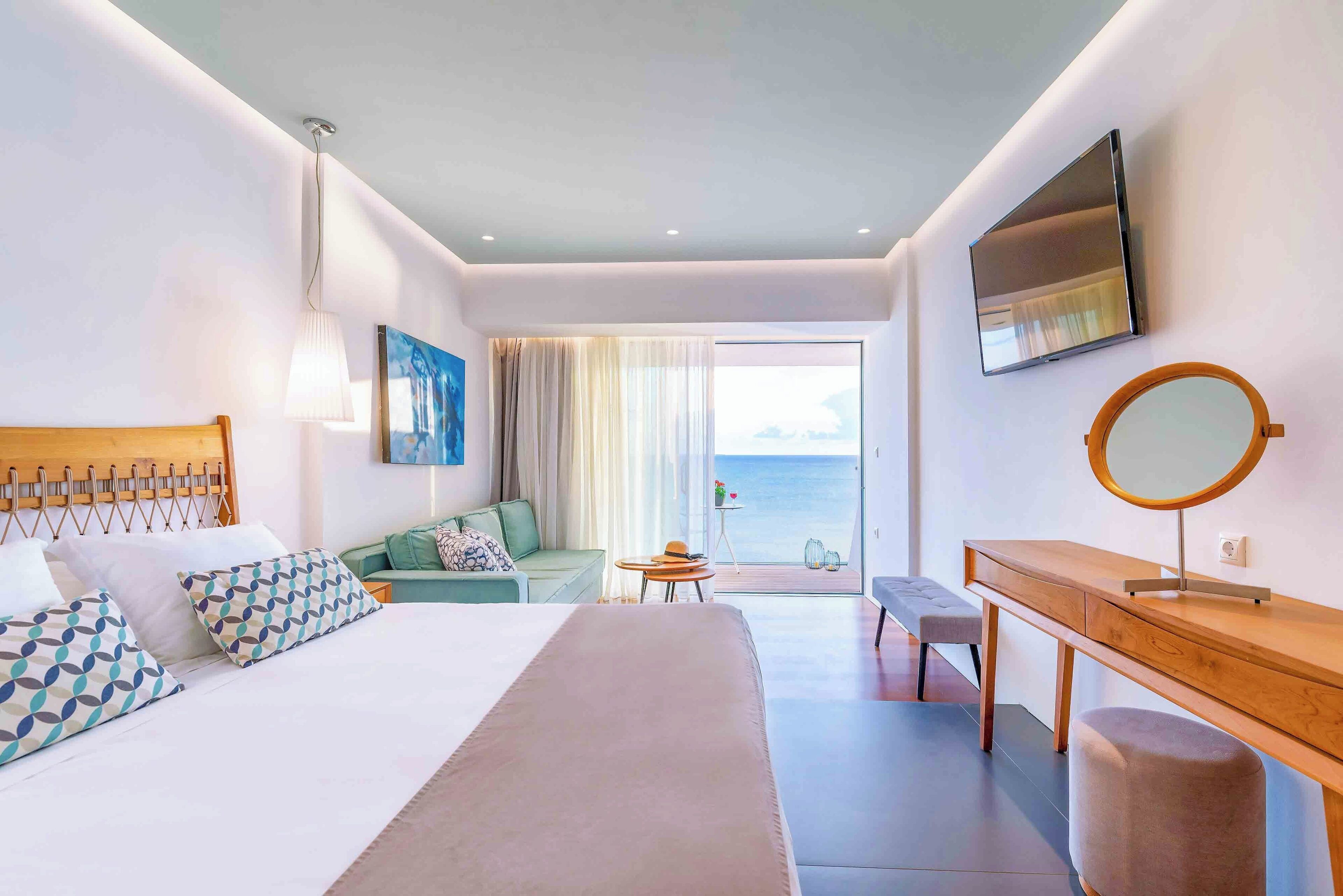 Ett modernt hotellrum med en stor säng, ett skrivbord i trä, en rund spegel och utsikt över havet genom fönstret.