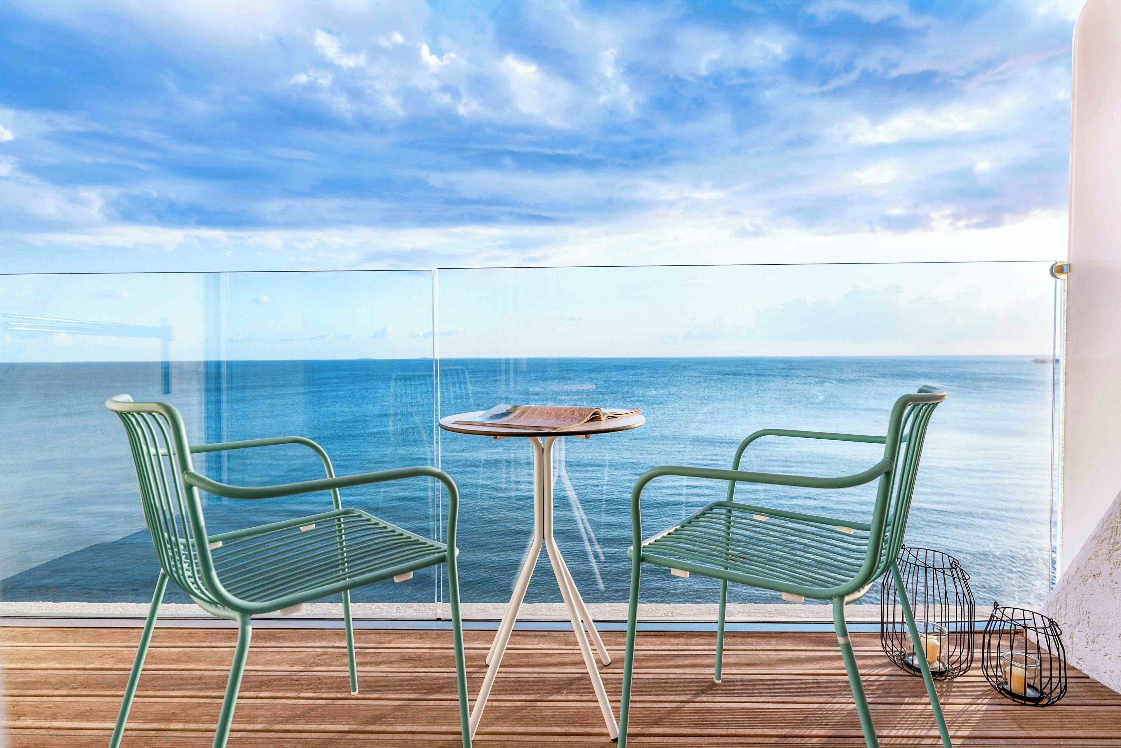A balcony with two green metal chairs and a small round table, overlooking a calm sea.