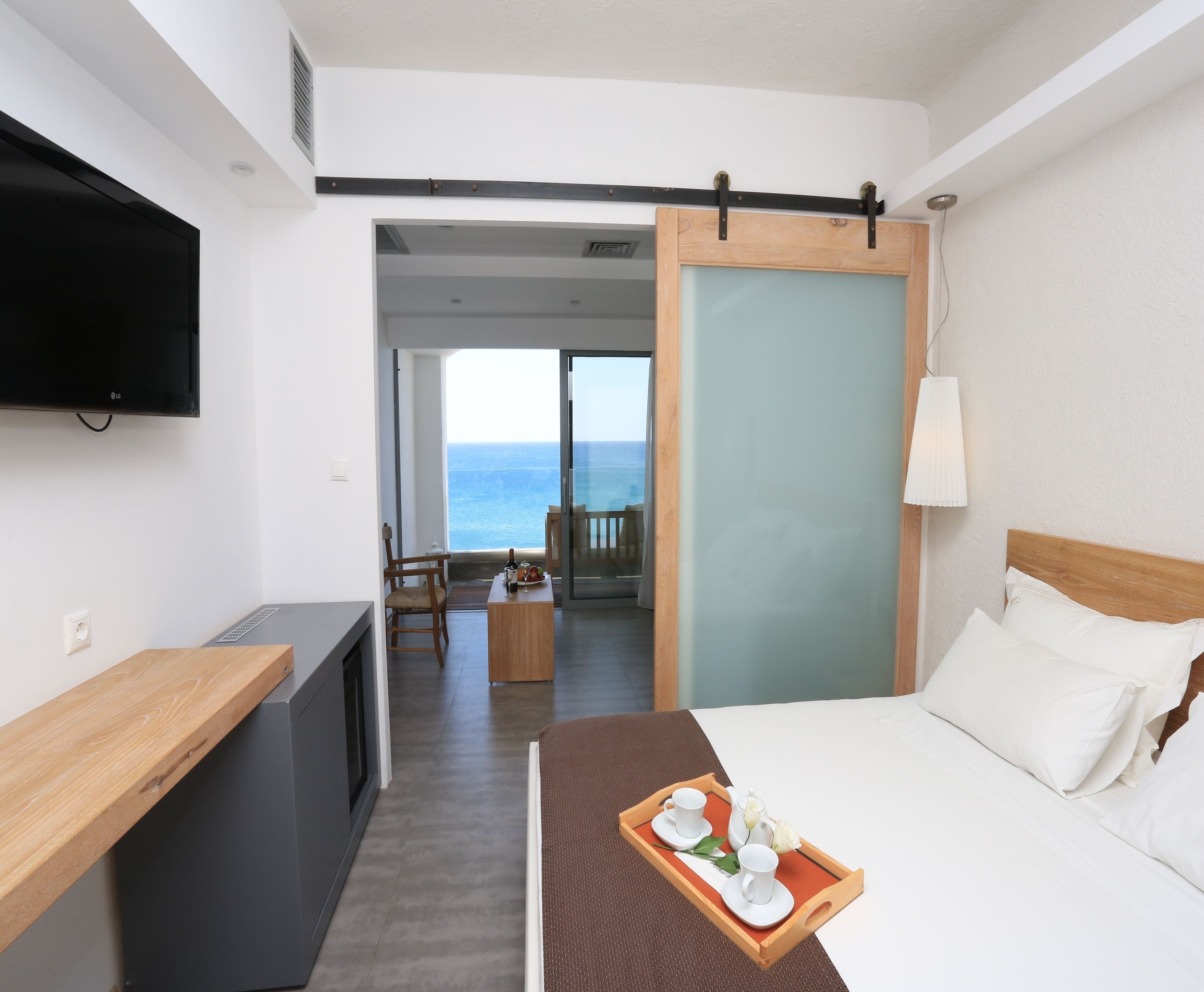 A hotel room with a bed, a desk, a TV, and a balcony with a view of the sea.
