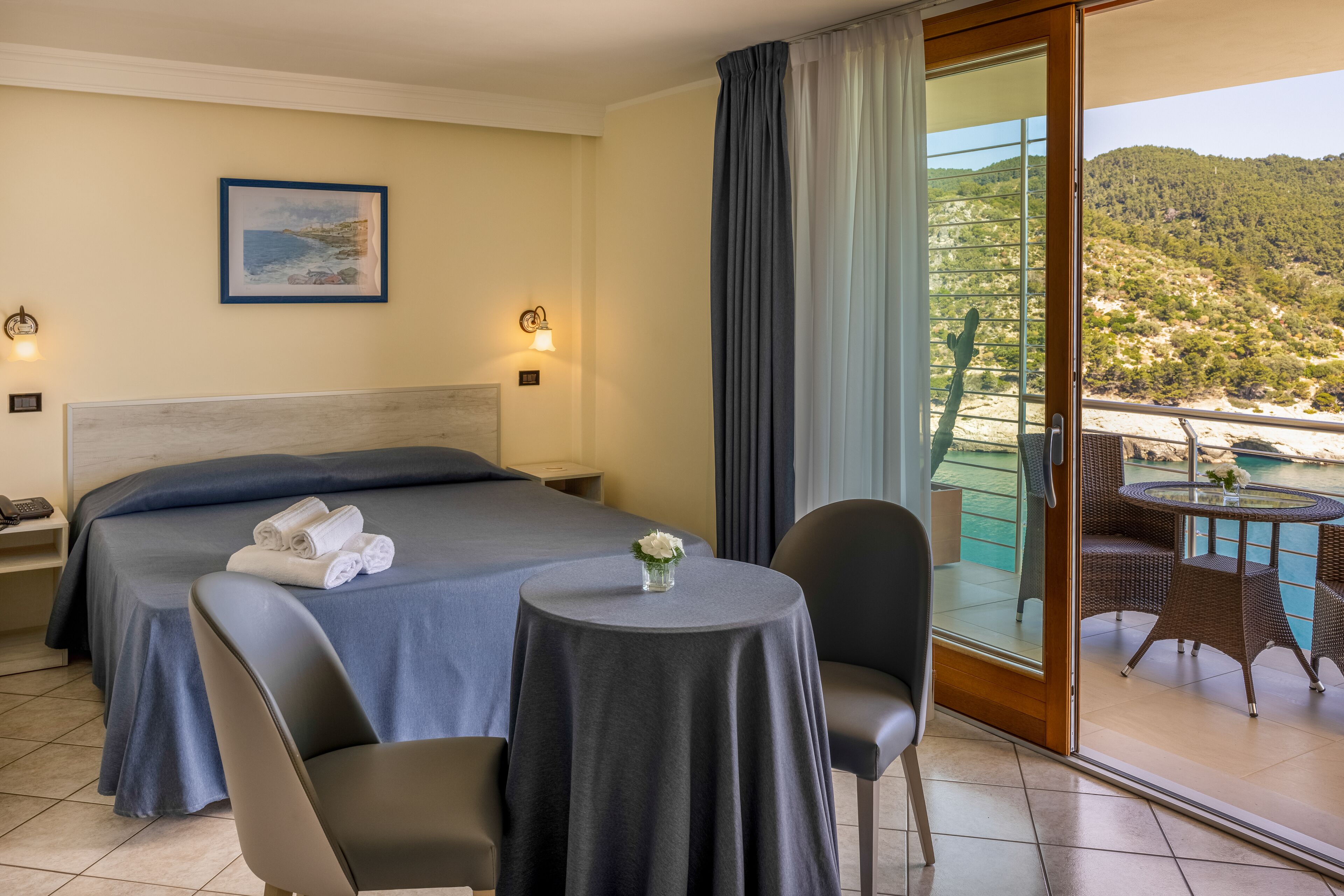 Una camera d'albergo con un letto, due comodini, un tavolino con un vaso, una sedia e un balcone con tavolo e sedie.