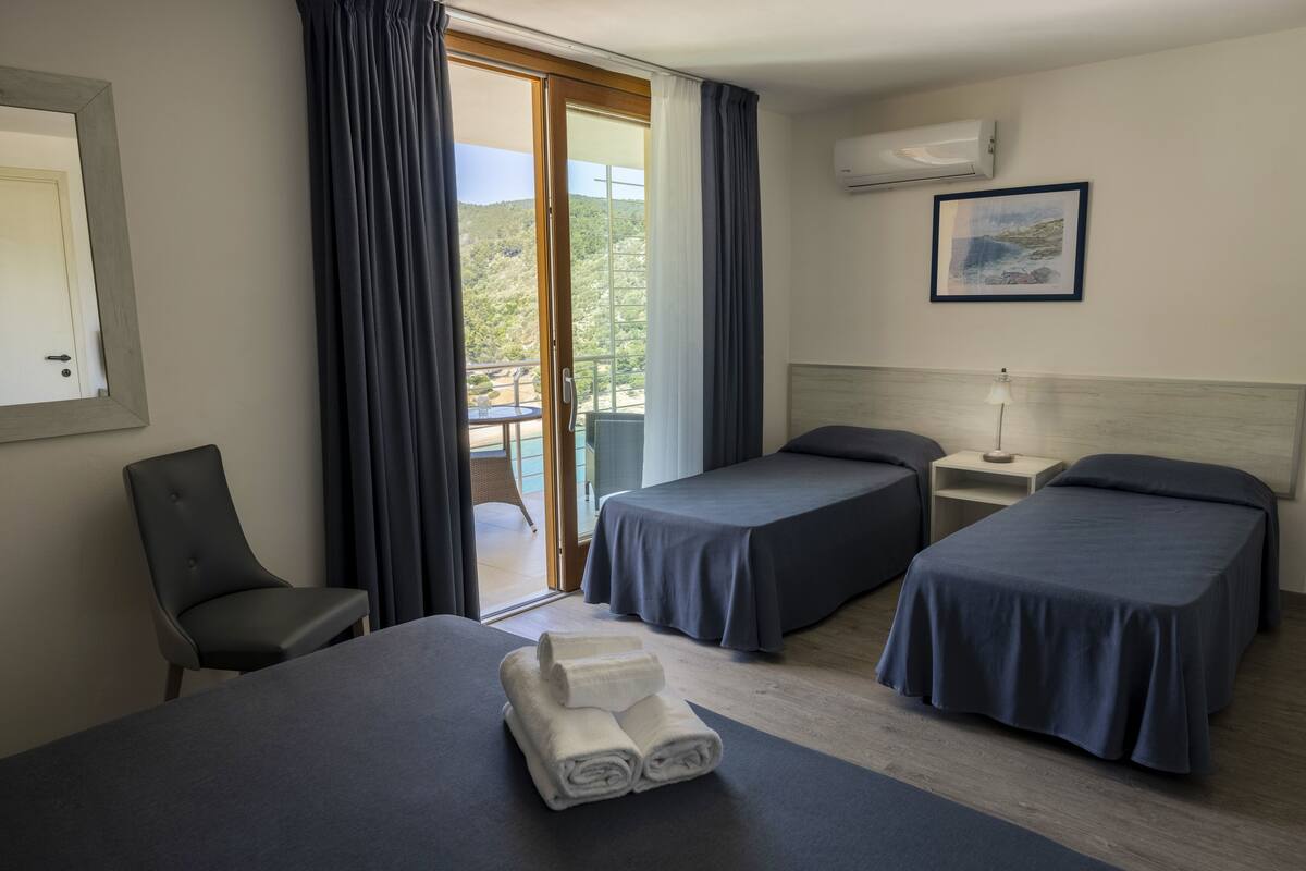 Ein Hotelzimmer mit zwei Betten, einem Sessel, einem Tisch mit Handtüchern, einem Nachttisch, einer Lampe und Blick ins Freie.