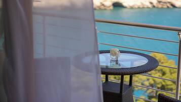 A balcony with a glass table and wicker chairs, overlooking a coastal area with a rocky shoreline.
