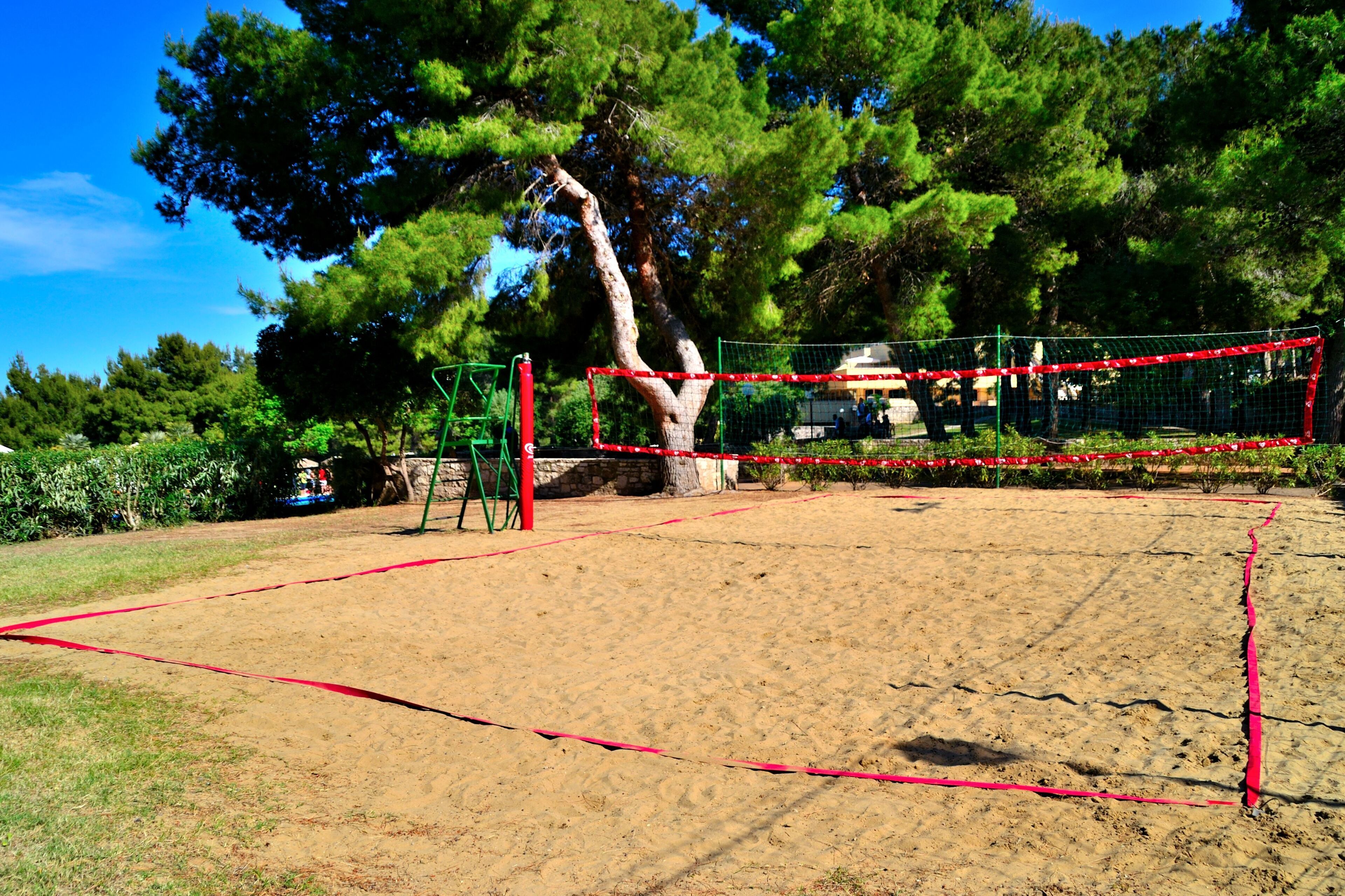 A sandy volleyball court with a net, surrounded by trees and a green fence.