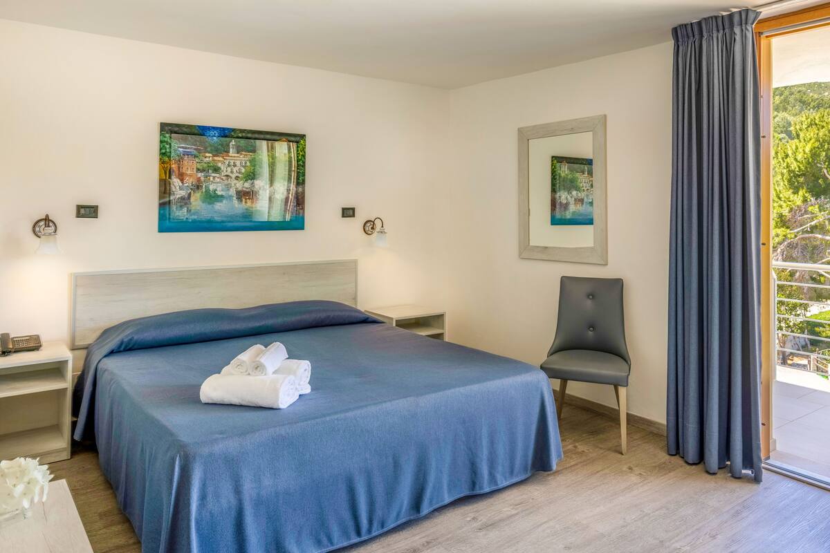 Ein Hotelzimmer mit Bett, Stuhl, Gemälde, Spiegel und Blick ins Freie.