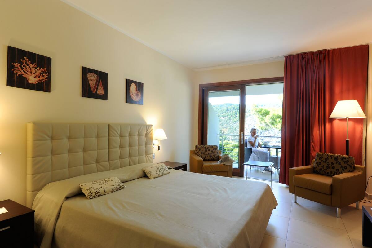 Ein Hotelzimmer mit einem großen Bett, zwei Sessel, einem Tisch und einem Balkon mit Aussicht.