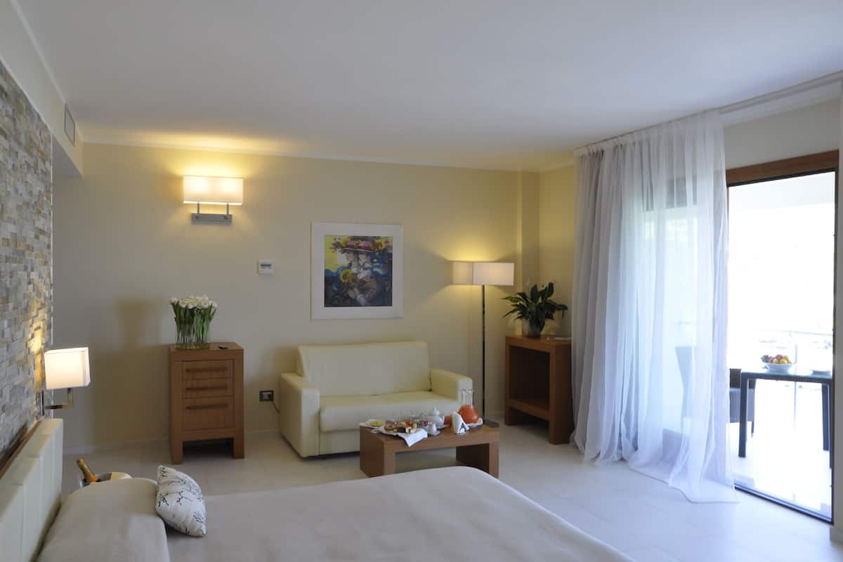 Ein Hotelzimmer mit Bett, Sofa, Couchtisch mit Frühstücksangeboten, Nachttisch mit Lampe, Wandbild und Fenster mit leichtem Vorhang.
