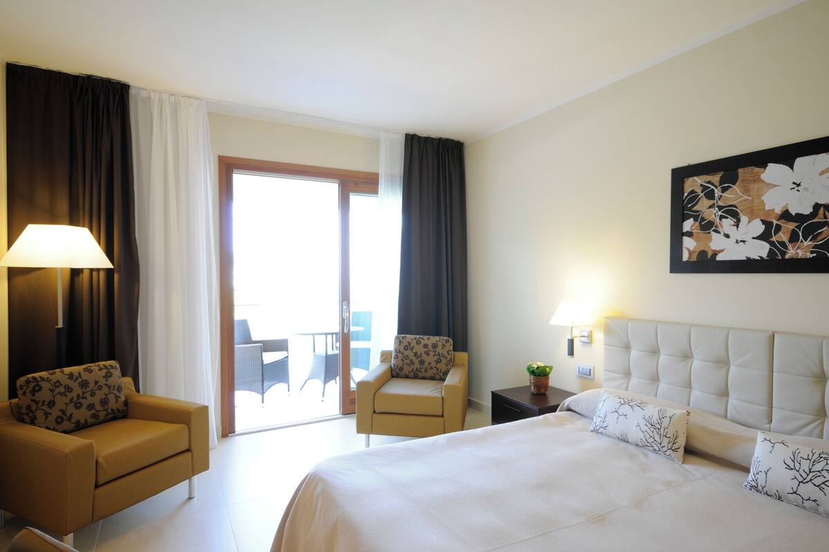 Ein Hotelzimmer mit einem Bett, zwei Sesseln, einem kleinen Tisch, einer Lampe und einem großen Fenster mit Vorhängen.