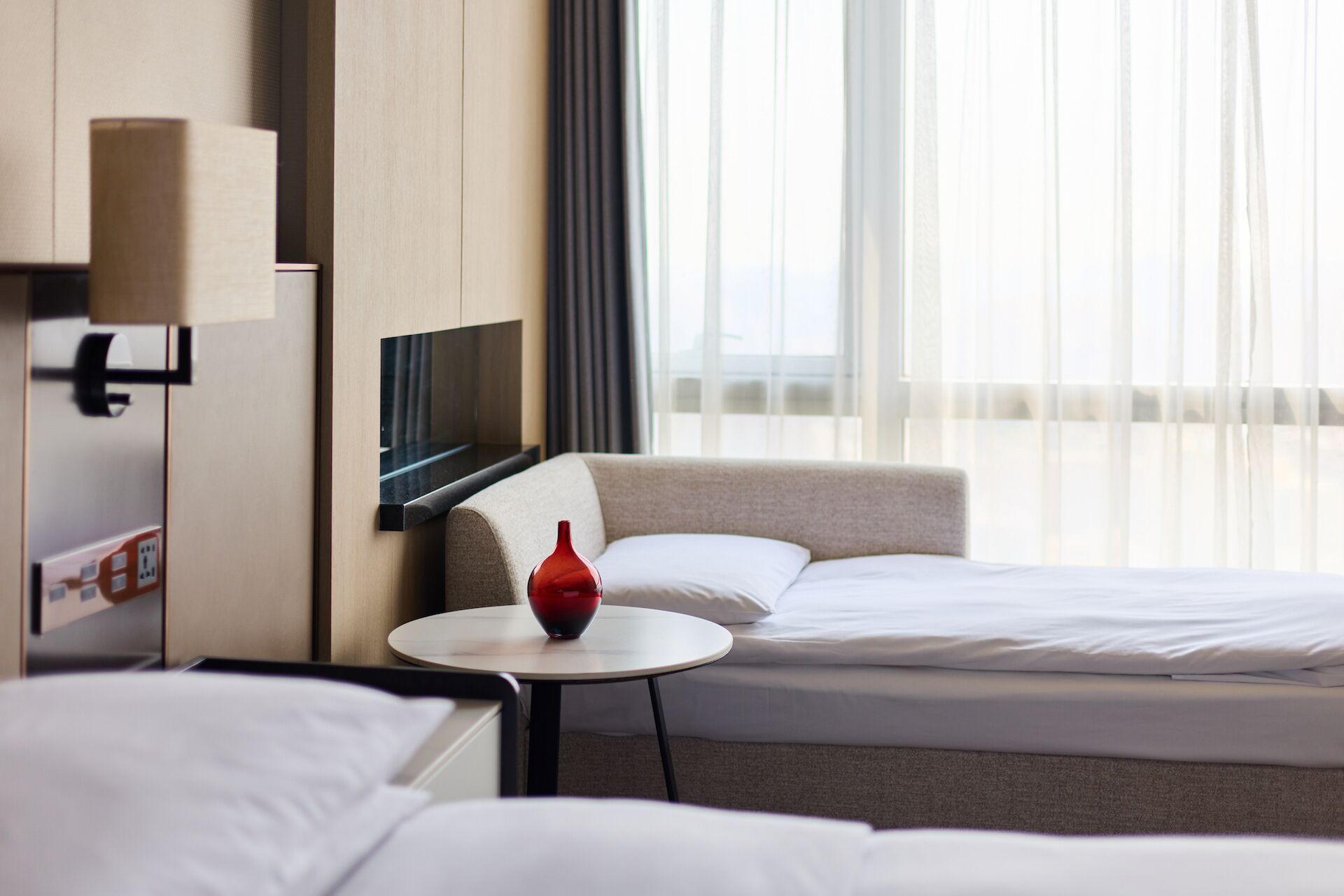 A hotel room with two beds, a small round table with a red vase, a flat-screen TV mounted on the wall, and a window with sheer curtains.
