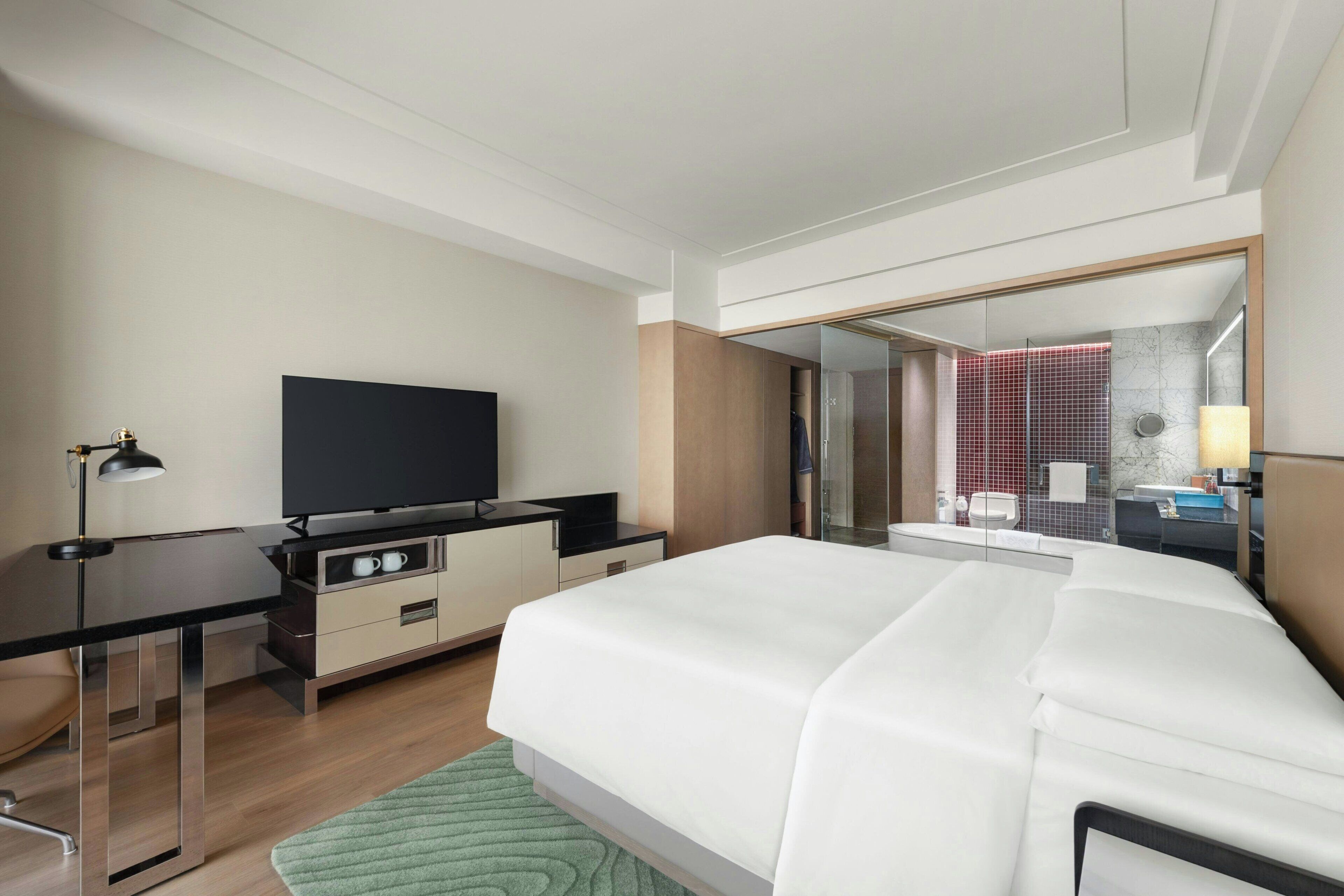 Ein modernes Hotelzimmer mit einem Bett, einem Schreibtisch mit Flachbildfernseher, einem Spiegel und einem durch eine Glastrennwand einsehbaren Badezimmer.