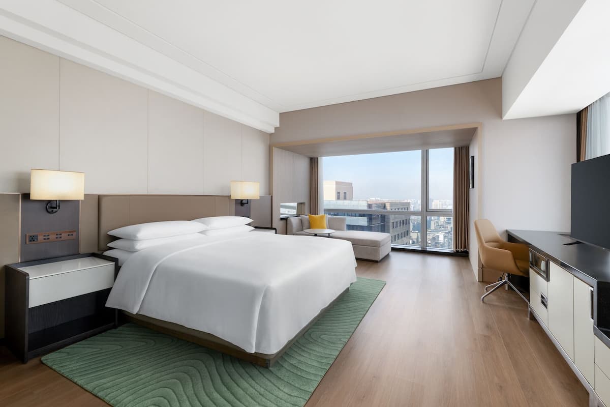 A hotel room with a bed, bedside tables, a desk, a chair, a television, and a large window with a city view.