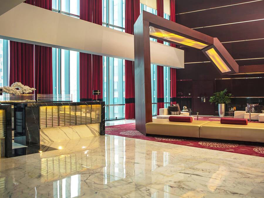 Un'ampia hall con pavimento in marmo, una reception, un'area salotto con divani e ampie finestre con tende rosse.