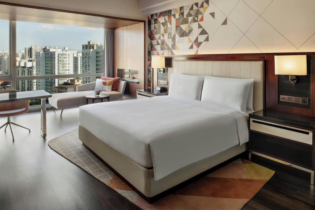 A modern hotel room with a large bed, a desk, a chair, and a view of the city skyline.