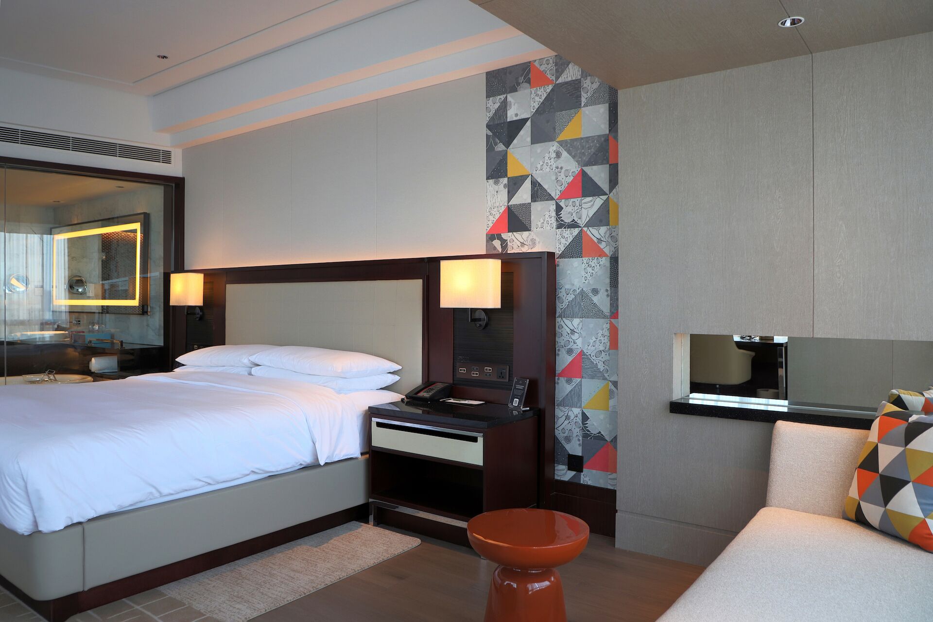 Una habitación de hotel con cama, mesita de noche, lámpara, televisor y un taburete pequeño.