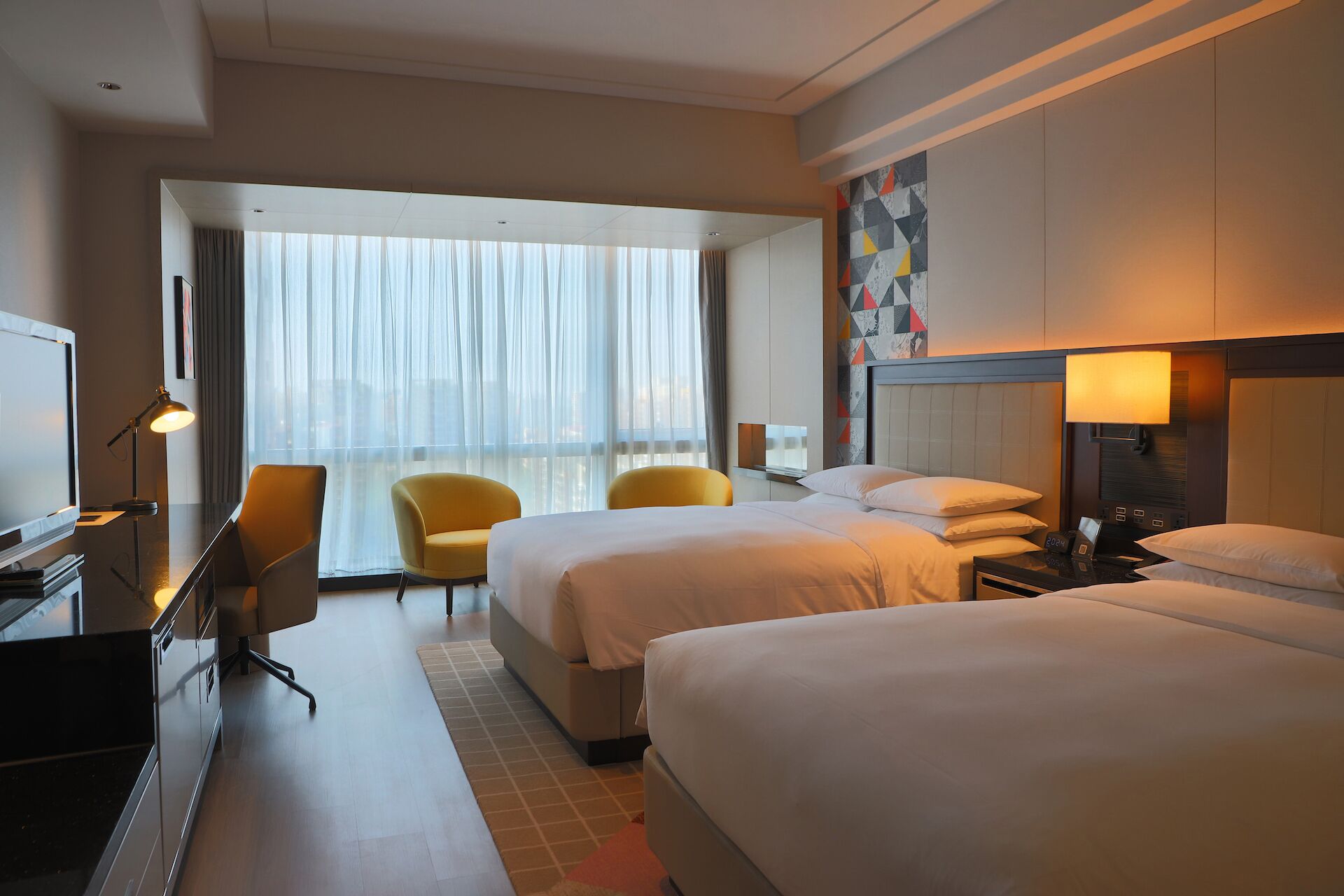 Habitación de hotel con dos camas, un escritorio con computadora, una silla y un amplio ventanal con cortinas.