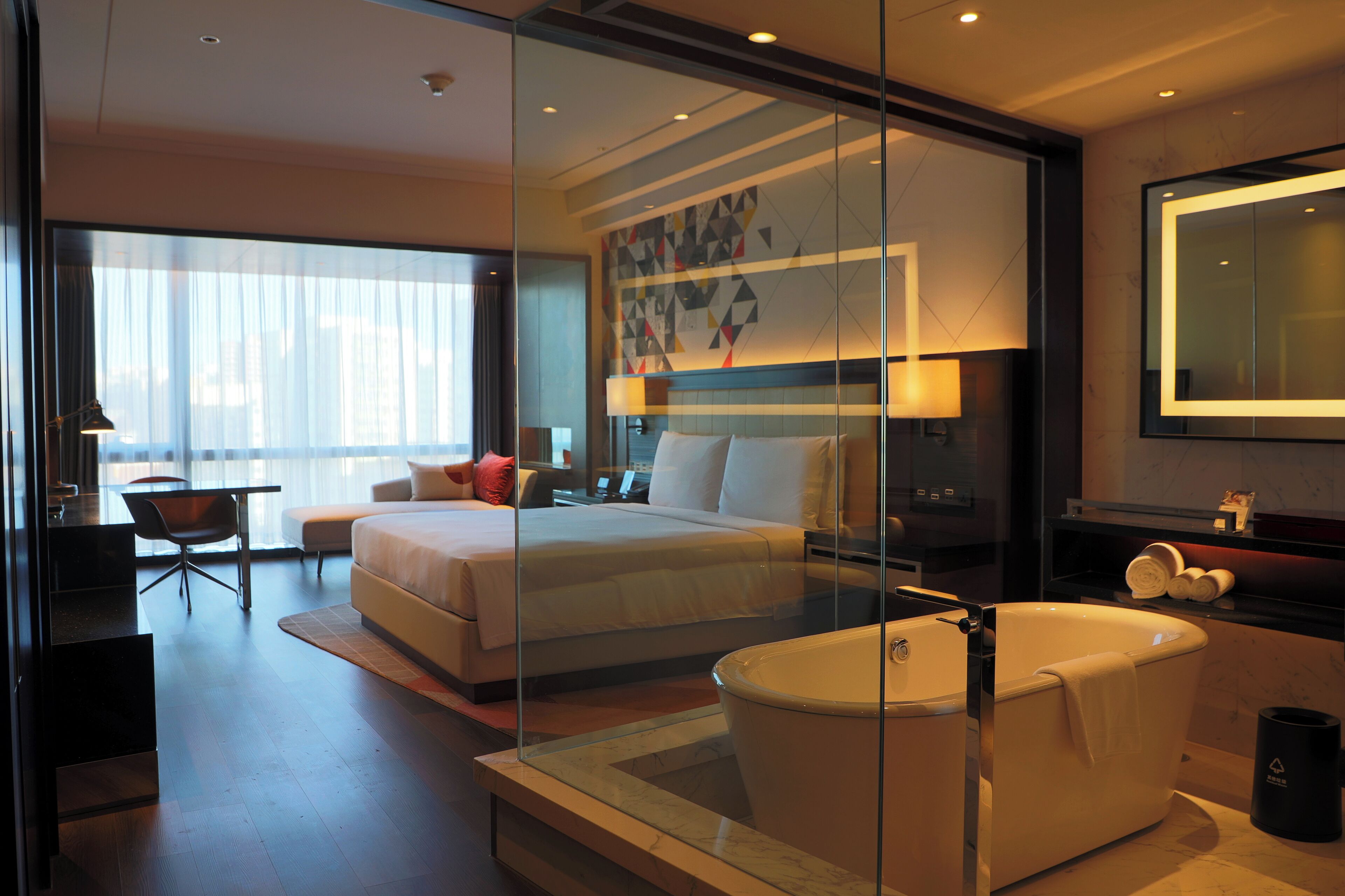 A modern hotel room with a large bed, a glass-enclosed bathtub, a desk, and a chair.