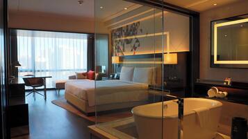 A modern hotel room with a large bed, a glass-enclosed bathtub, a desk, and a chair.