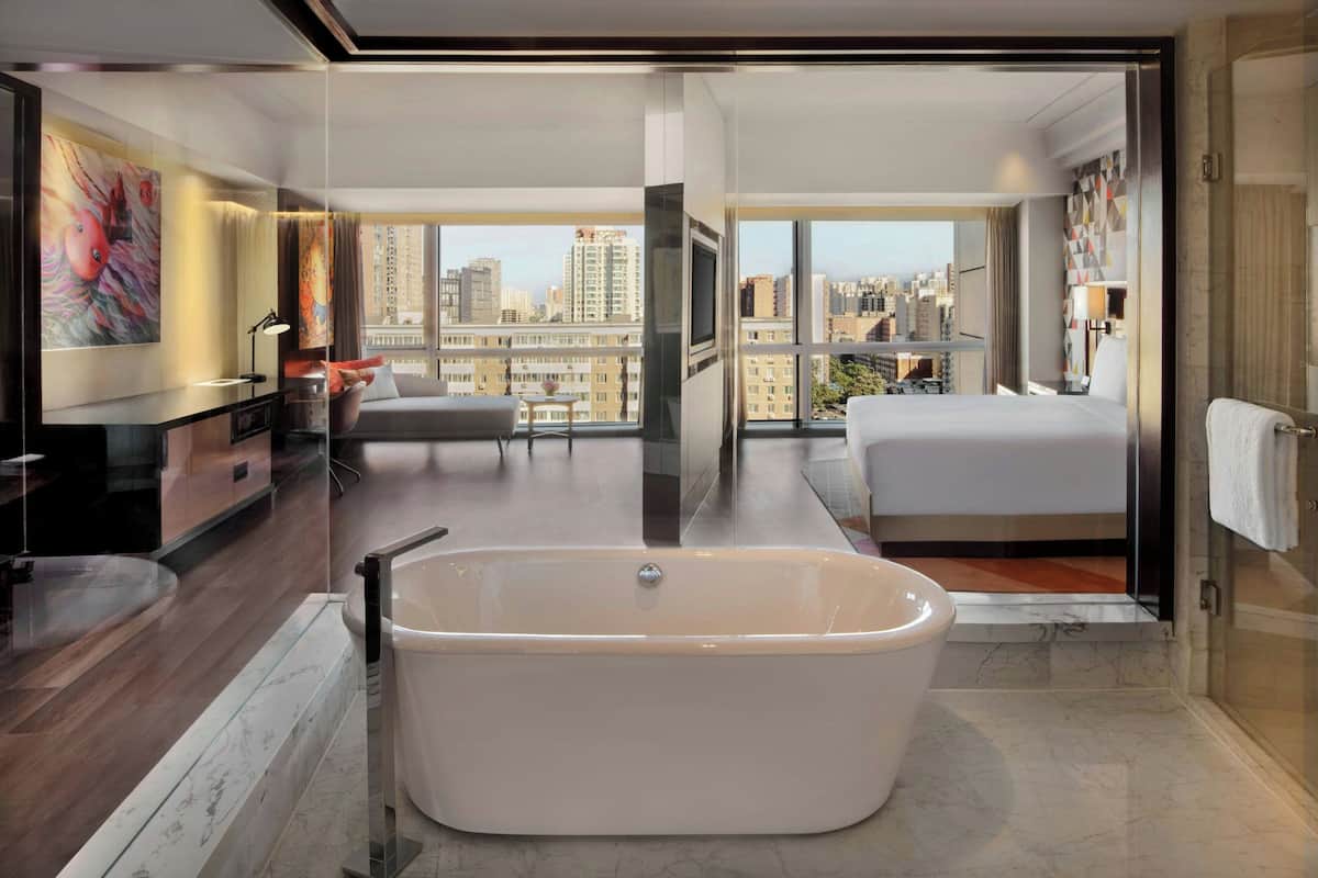 A modern hotel room with a large bathtub, a bed, a desk, and a city view.