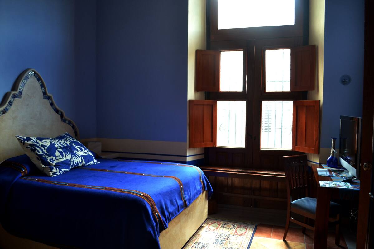 A bedroom with a bed, a desk, and a window with shutters.