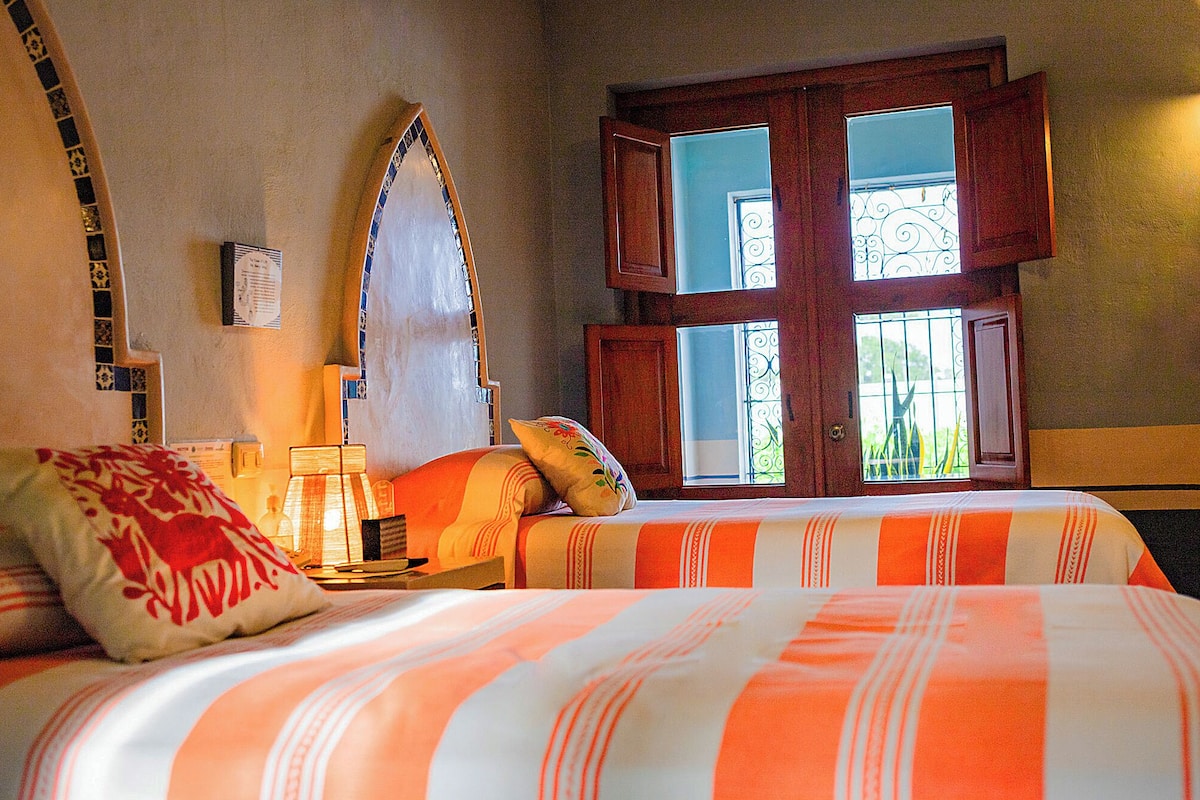 A bedroom with two beds, a nightstand, a lamp, and a window with decorative iron bars.
