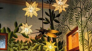 Una habitación con un mural de follaje tropical, un sofá de madera con cojines estampados, una mesa de centro de madera y un colgante de techo con forma de estrella.