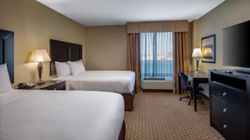 A hotel room with two beds, a desk, a chair, a TV, and an air conditioning unit.
