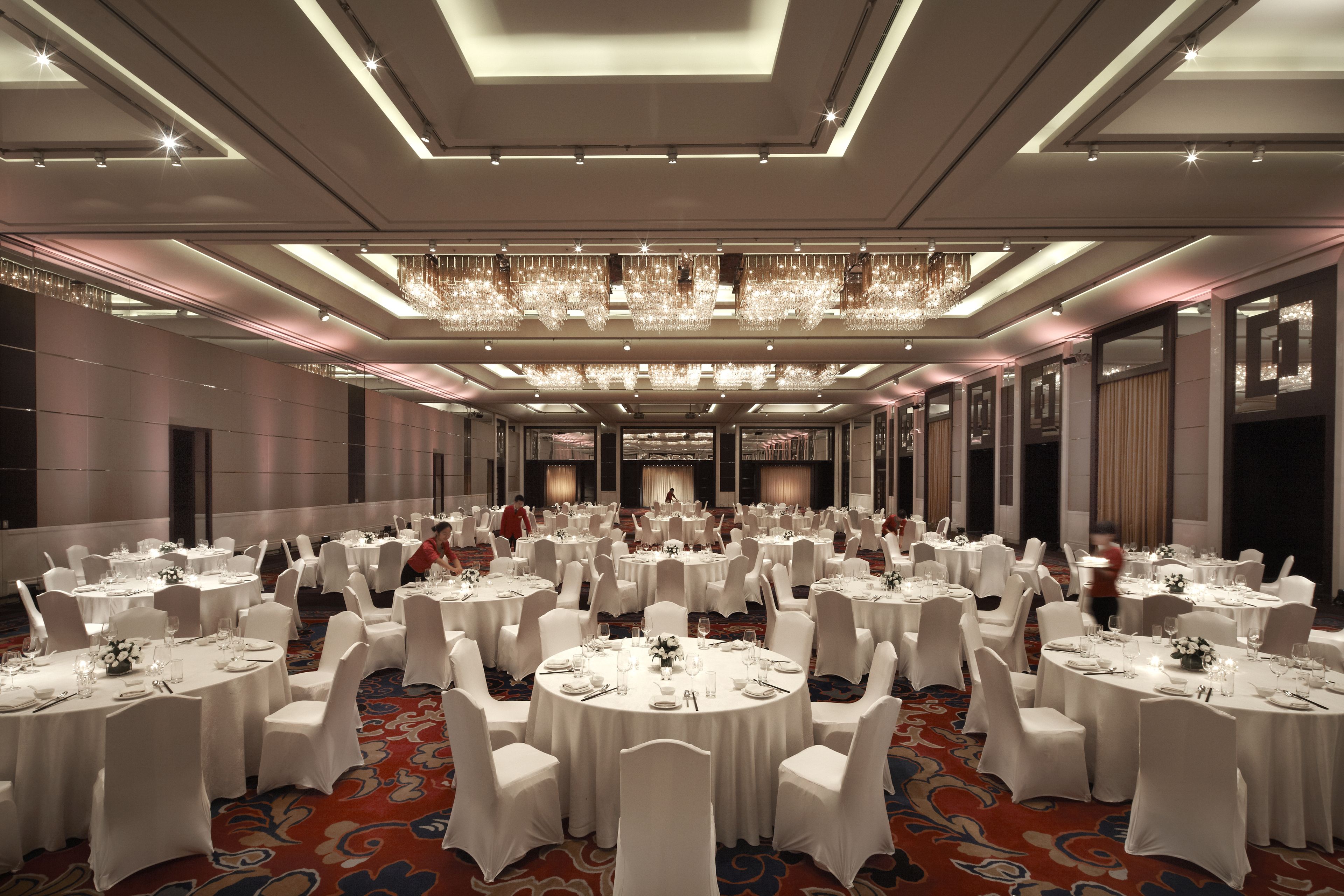 Un salón de banquetes con mesas redondas, sillas blancas, candelabros y una alfombra roja.
