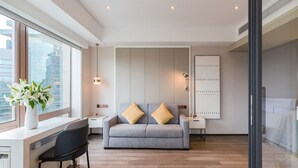 A modern living room with a grey sofa, two cushions, a side table, a desk with a chair, a vase with flowers, and a large window. - Beijing Pudi Hotel (Beijing)