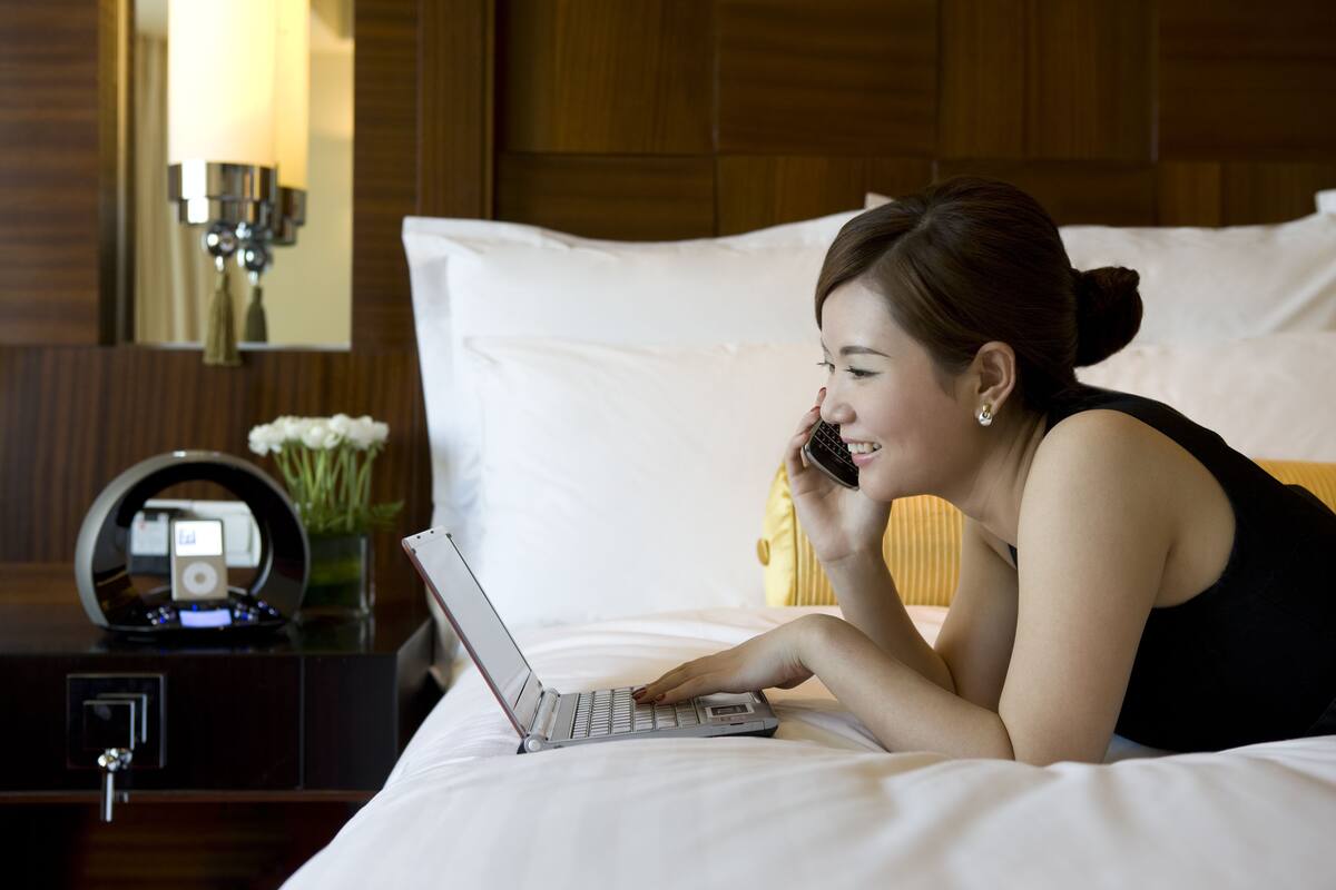 A person using a laptop on a bed in a hotel room with a wooden headboard, a bedside table with a lamp, and a small vase with flowers.
