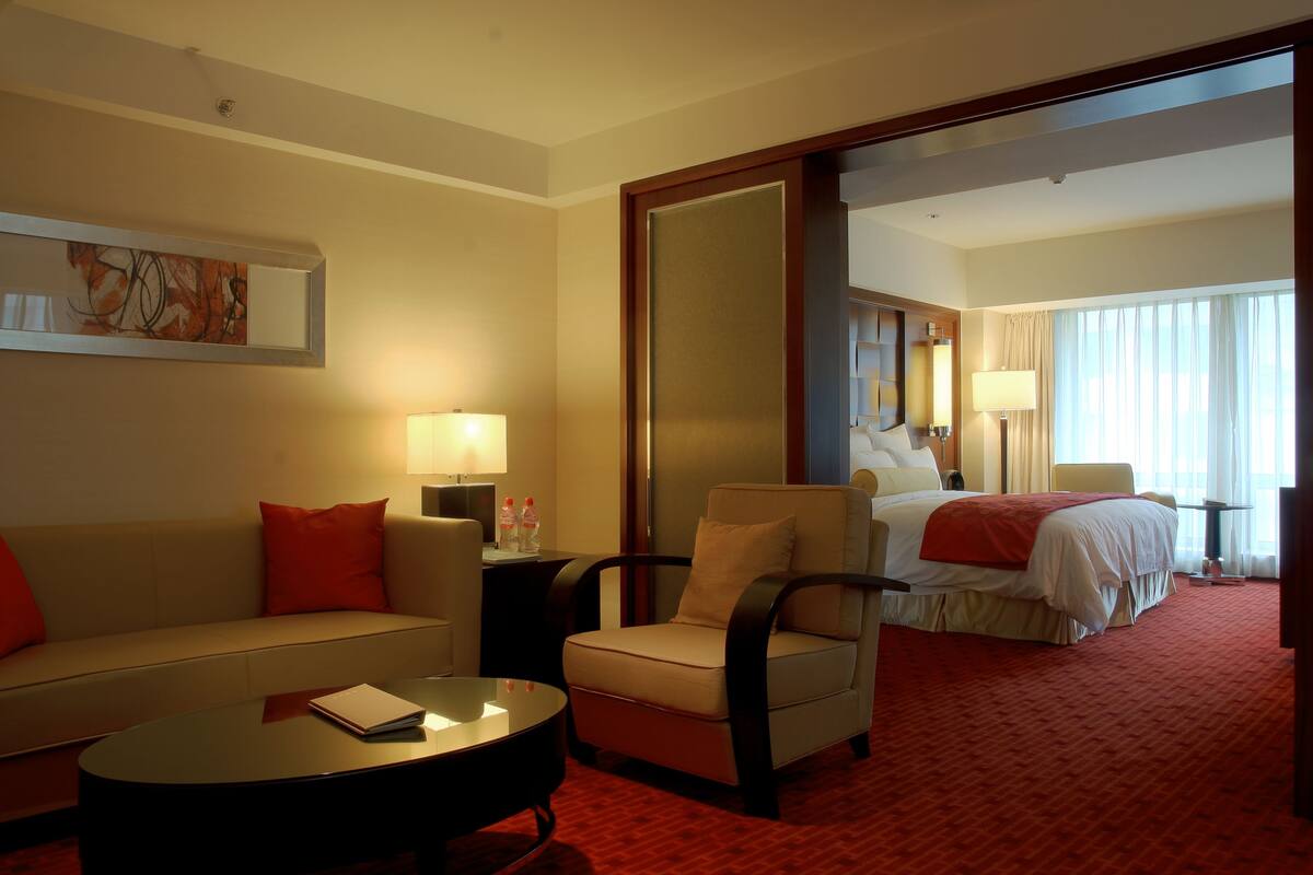 A hotel room with a sofa, armchair, and a bed. There is a lamp on a side table and a painting on the wall.