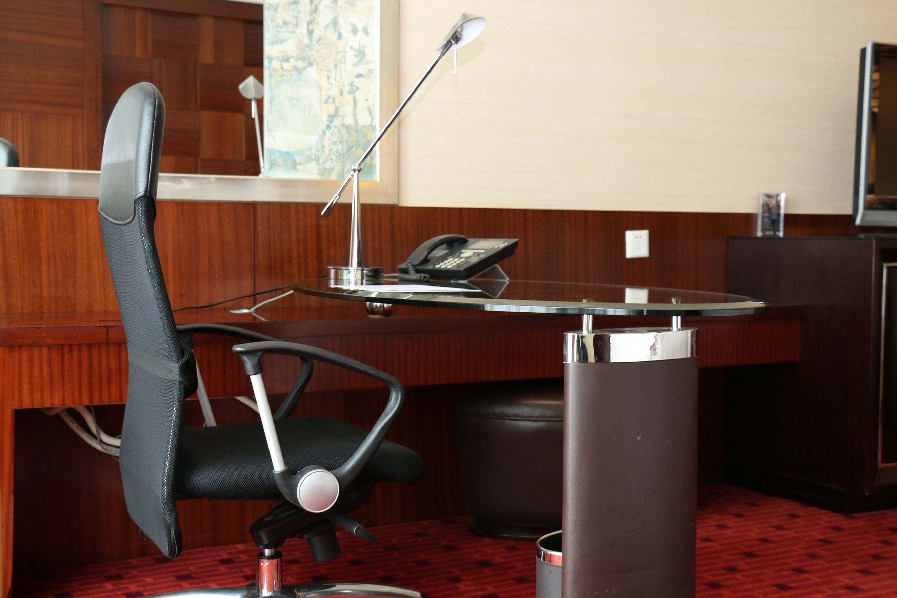 Una habitación de hotel con un escritorio de madera, una silla de oficina negra, un teléfono y una lámpara.