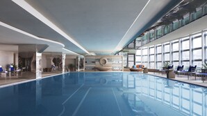 A large indoor swimming pool with clear blue water, surrounded by lounge chairs and tables, and large windows providing natural light. - Beijing Pudi Hotel (Beijing)