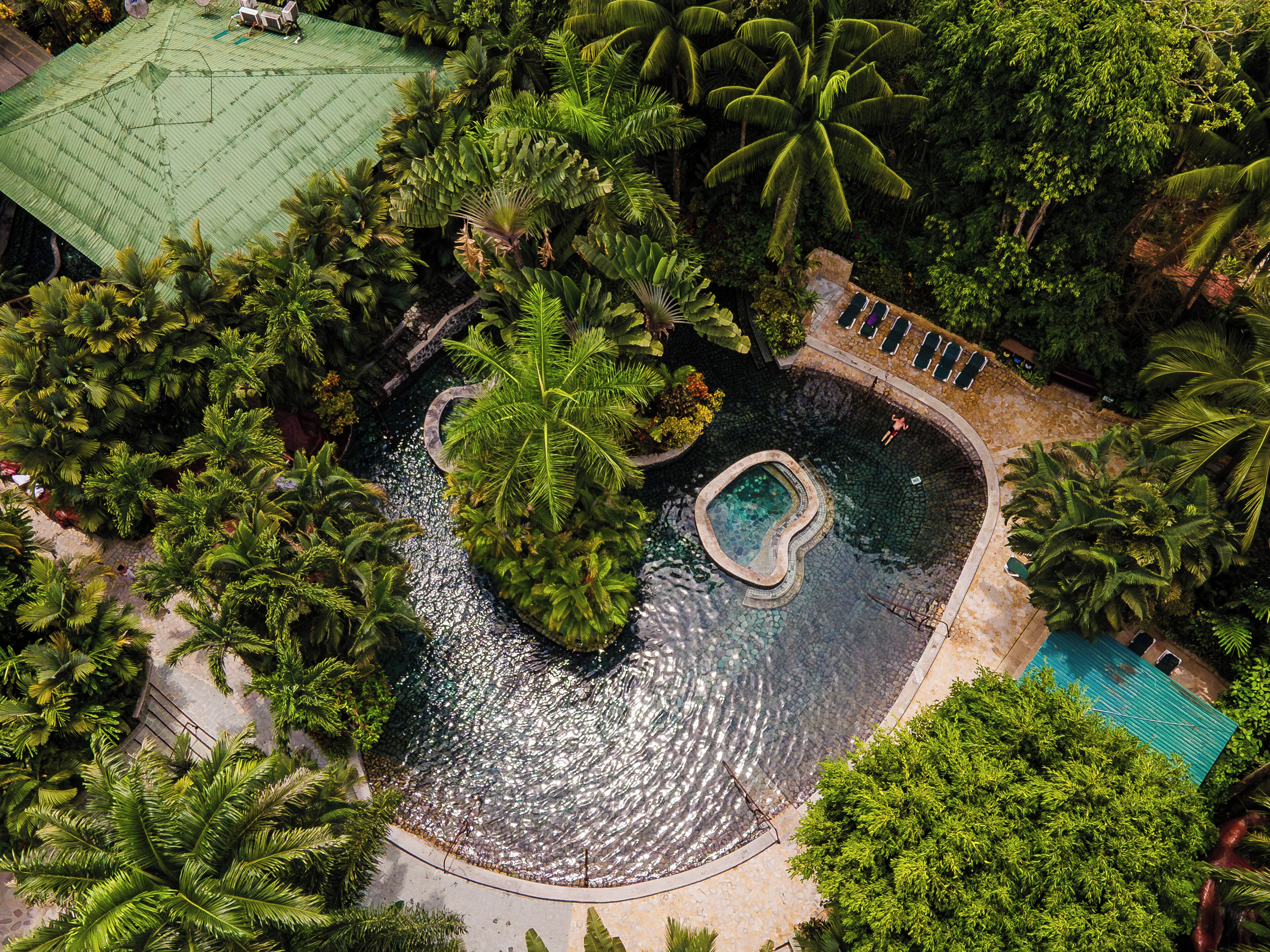 An aerial view of a resort with a central swimming pool surrounded by lush greenery and palm trees.