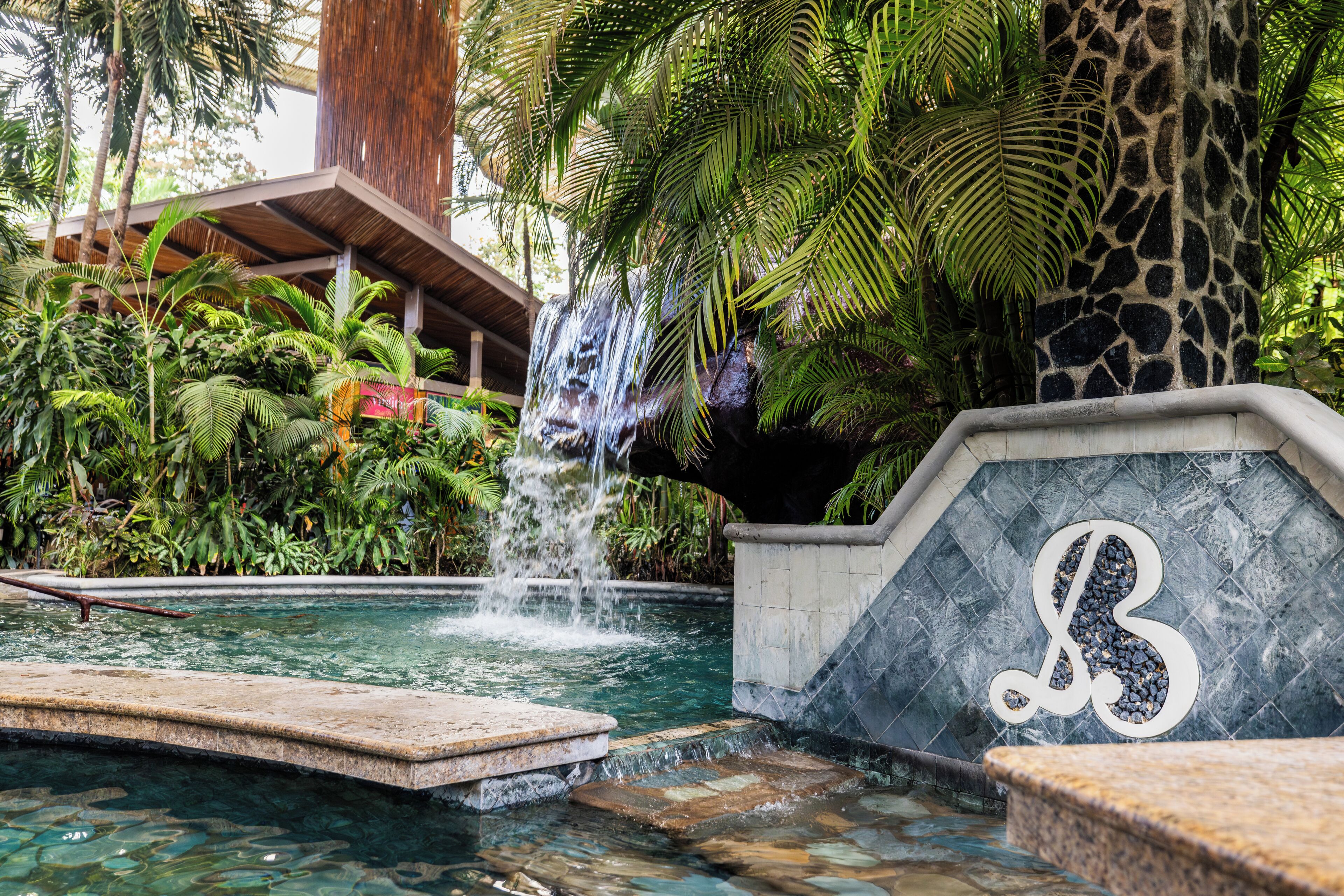 A pool with a waterfall, surrounded by lush greenery and a stone wall with a monogram.