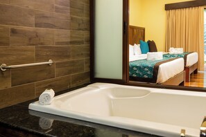 A hotel bathroom with a bathtub, a towel rack, and a mirror reflecting a room with a bed and curtains.