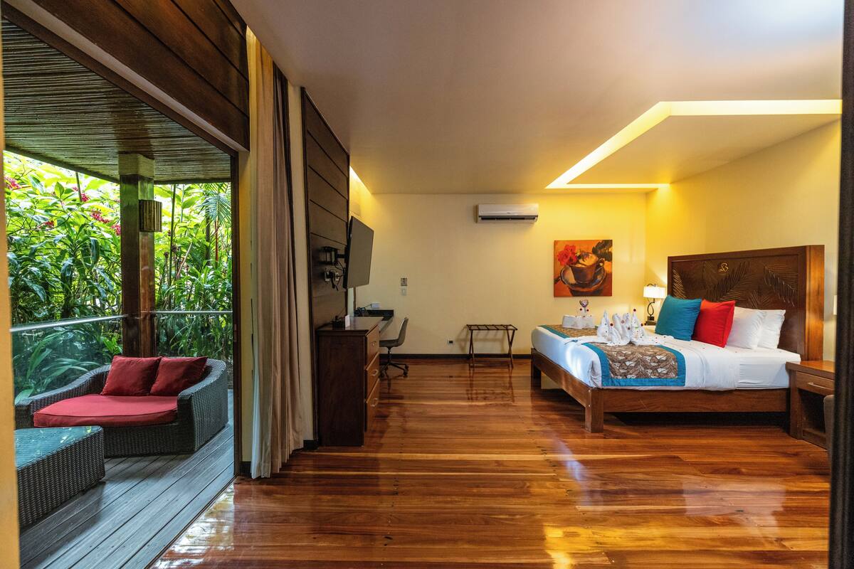 Ein Hotelzimmer mit Holzboden, einem Bett mit bunten Kissen, einem Schreibtisch mit Stuhl und einem Balkon mit Korbmöbeln.