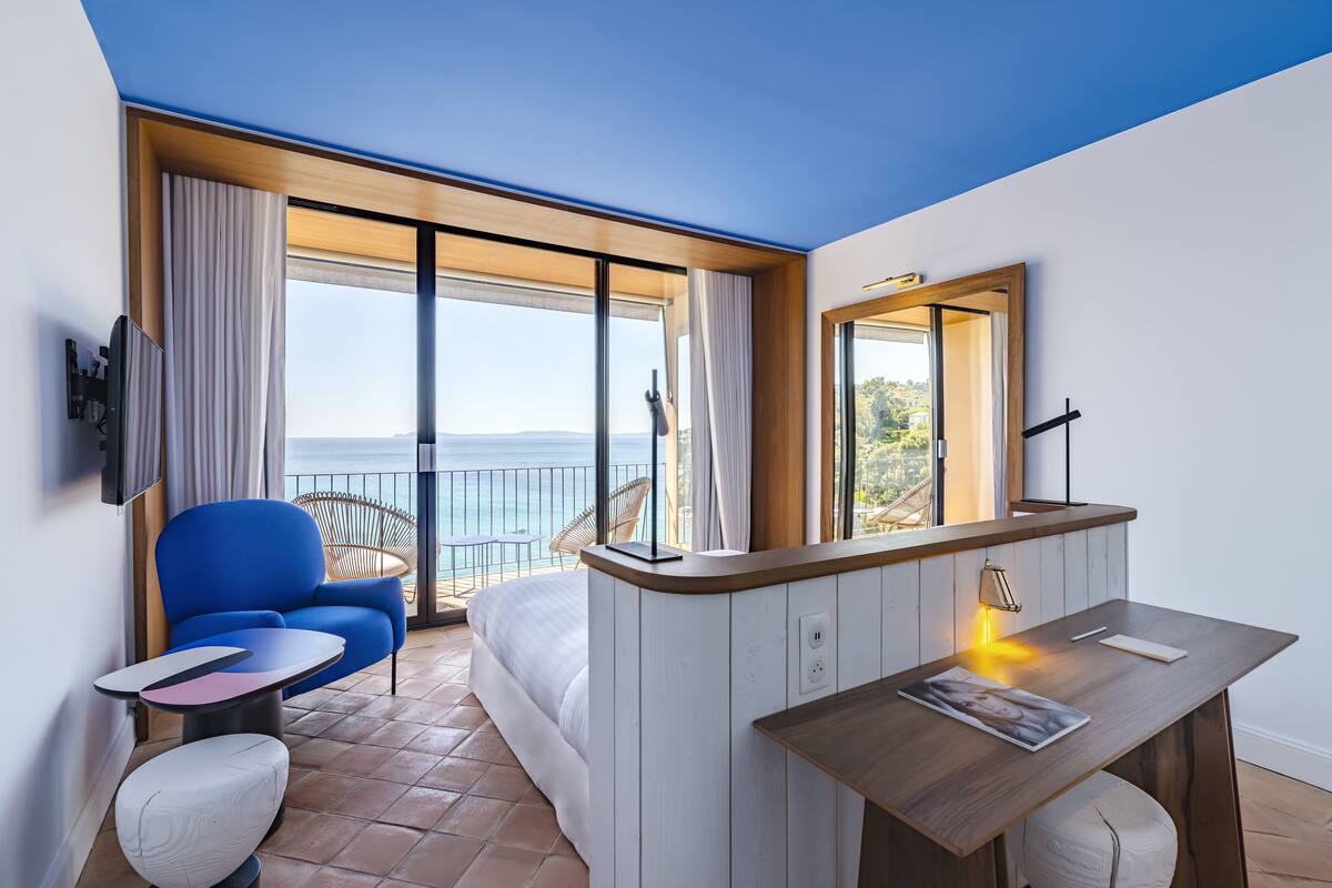 Una habitación de hotel moderna con una cama grande, un sillón azul, una mesita pequeña, un escritorio de madera y un ventanal con vistas al mar.