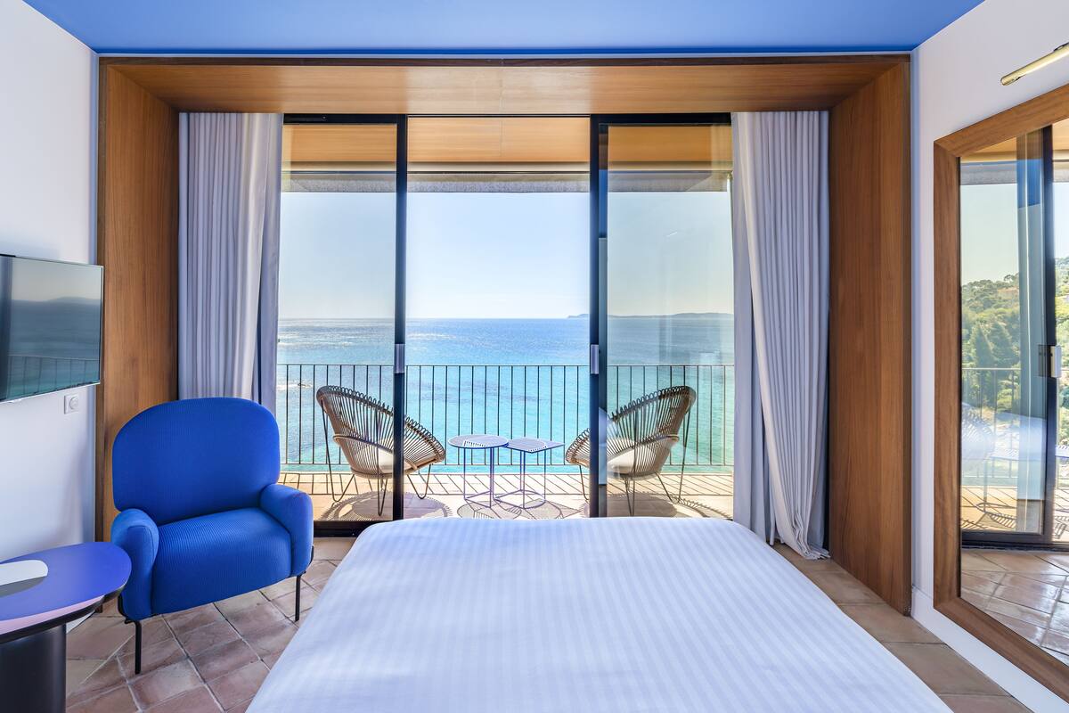 Habitación de hotel con una cama grande, televisión, una silla azul, una mesita y un balcón con vista al mar.