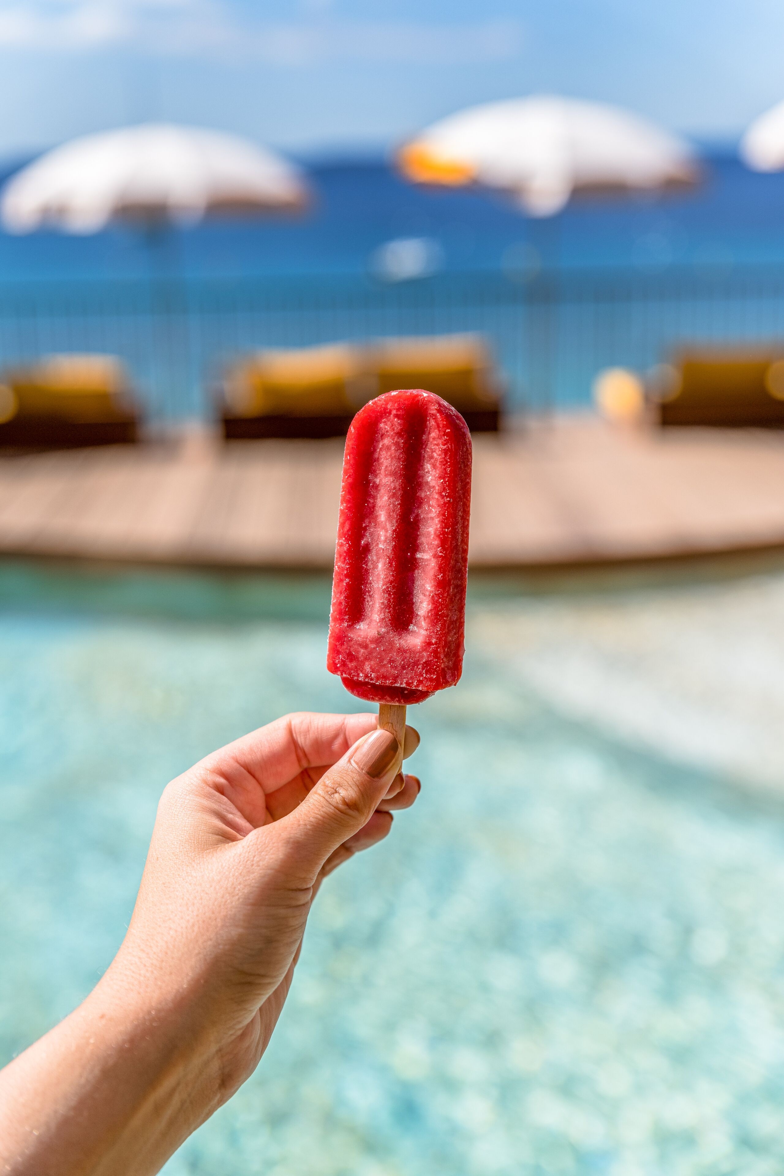 Una mano che tiene un ghiacciolo rosso sopra una piscina con ombrelloni sullo sfondo.