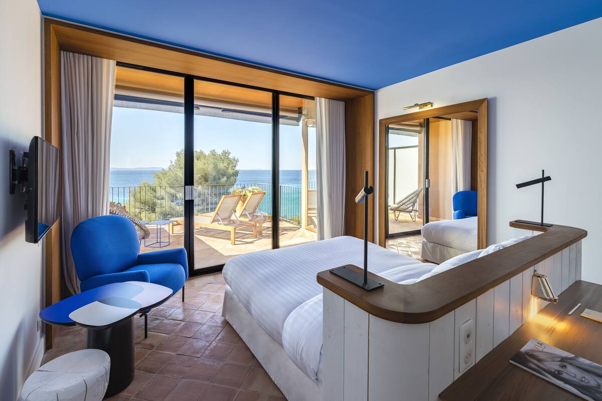 Una habitación de hotel moderna con una cama grande, un rincón de descanso con una silla azul, una mesita y vistas al mar a través de amplias ventanas.