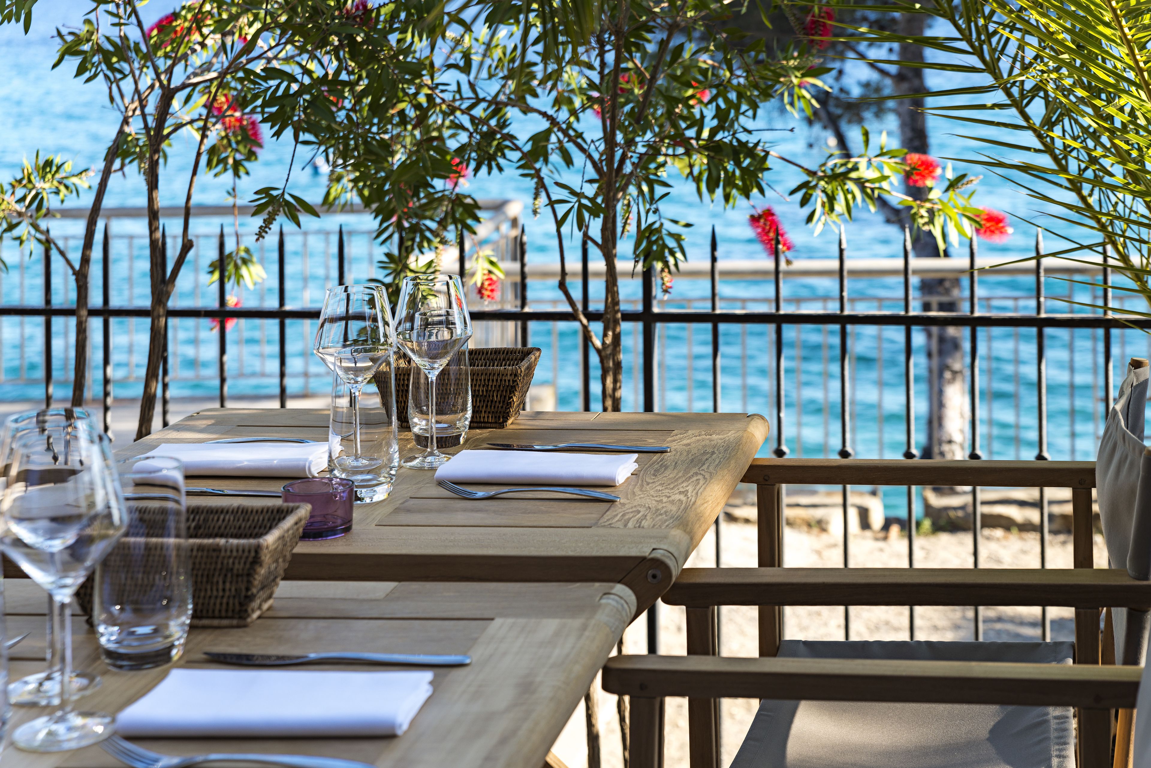 A wooden table set for dining with glasses and a basket, overlooking a beach with a railing and trees.