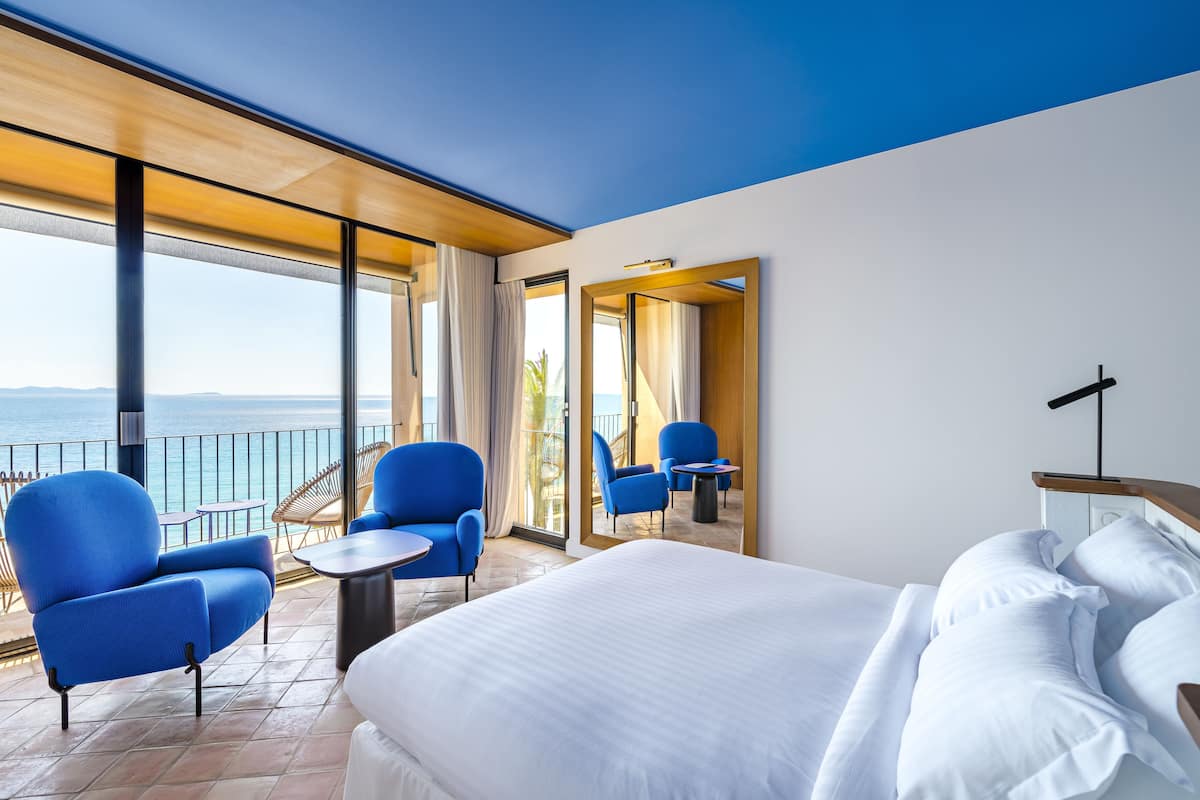Habitación de hotel con una cama grande, dos sillones azules, una mesita y un balcón con vistas al mar.