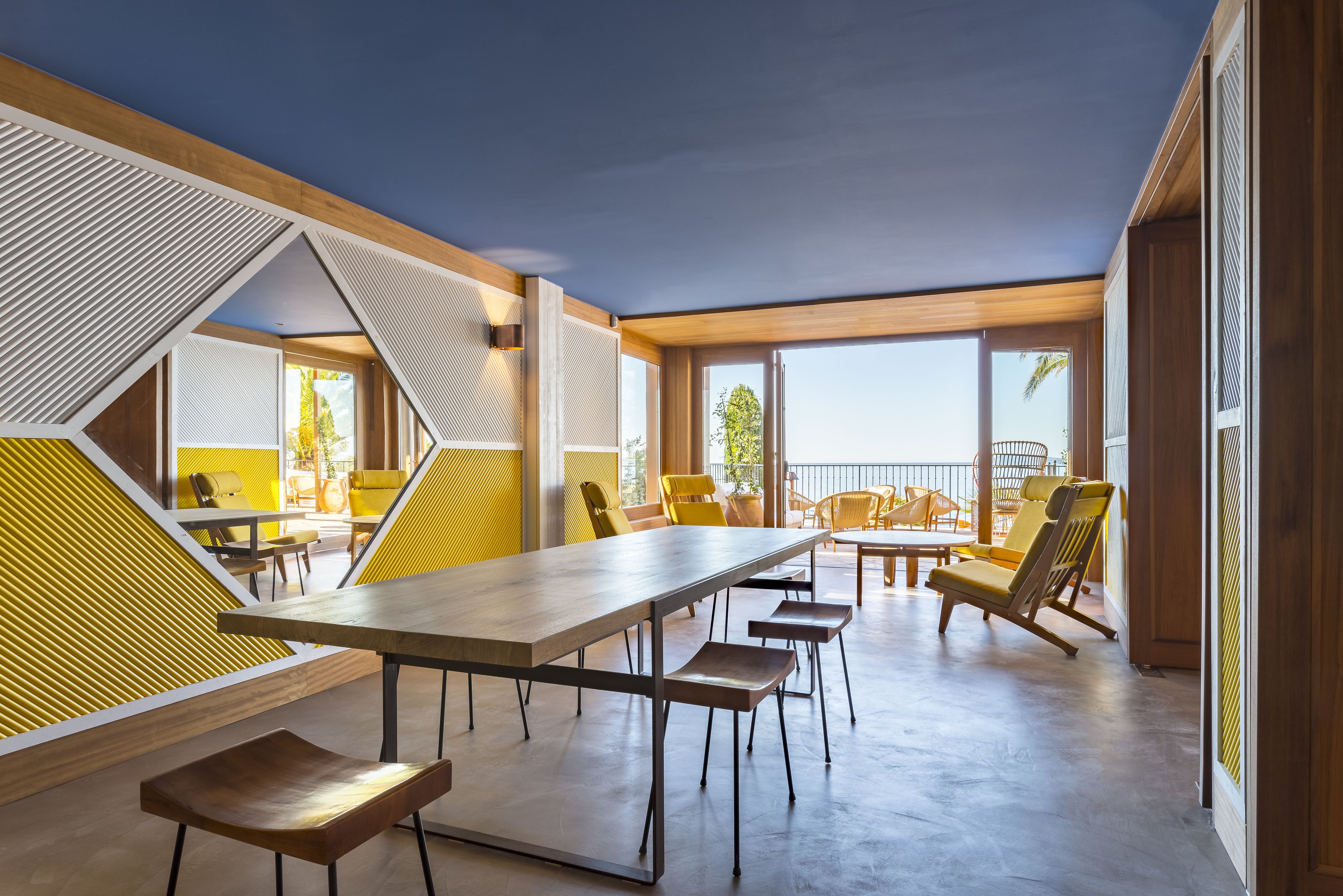 A modern dining area with a long table and multiple chairs, large windows, and a view of the outdoors.