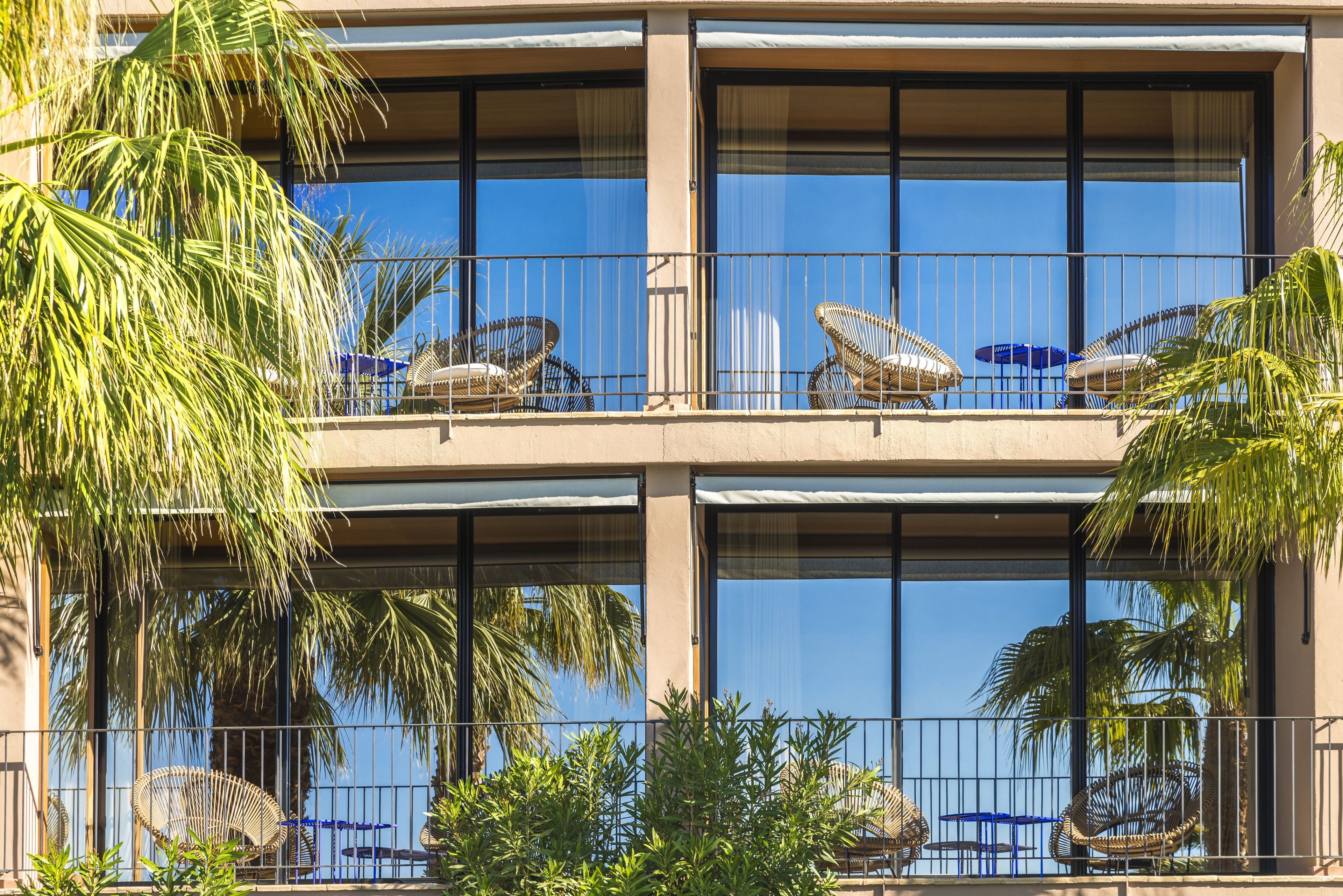 A hotel with a balcony featuring wicker chairs and umbrellas, surrounded by palm trees.
