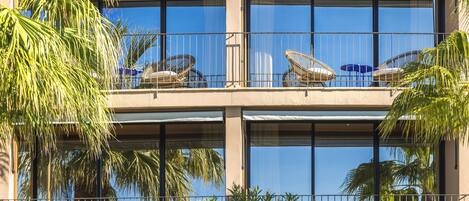 A hotel with a balcony featuring wicker chairs and umbrellas, surrounded by palm trees.