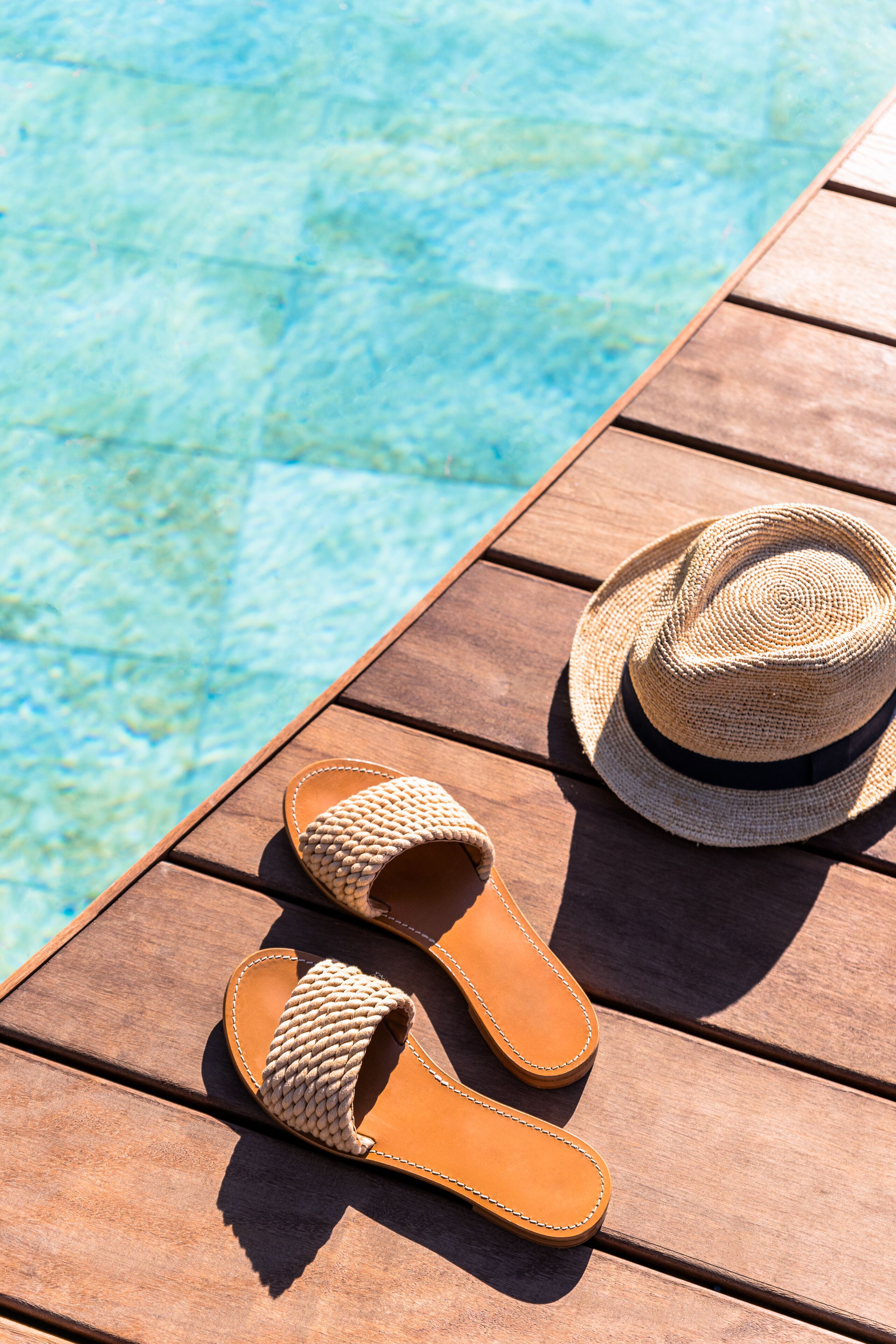 A pair of woven sandals and a straw hat on a wooden table by a pool.