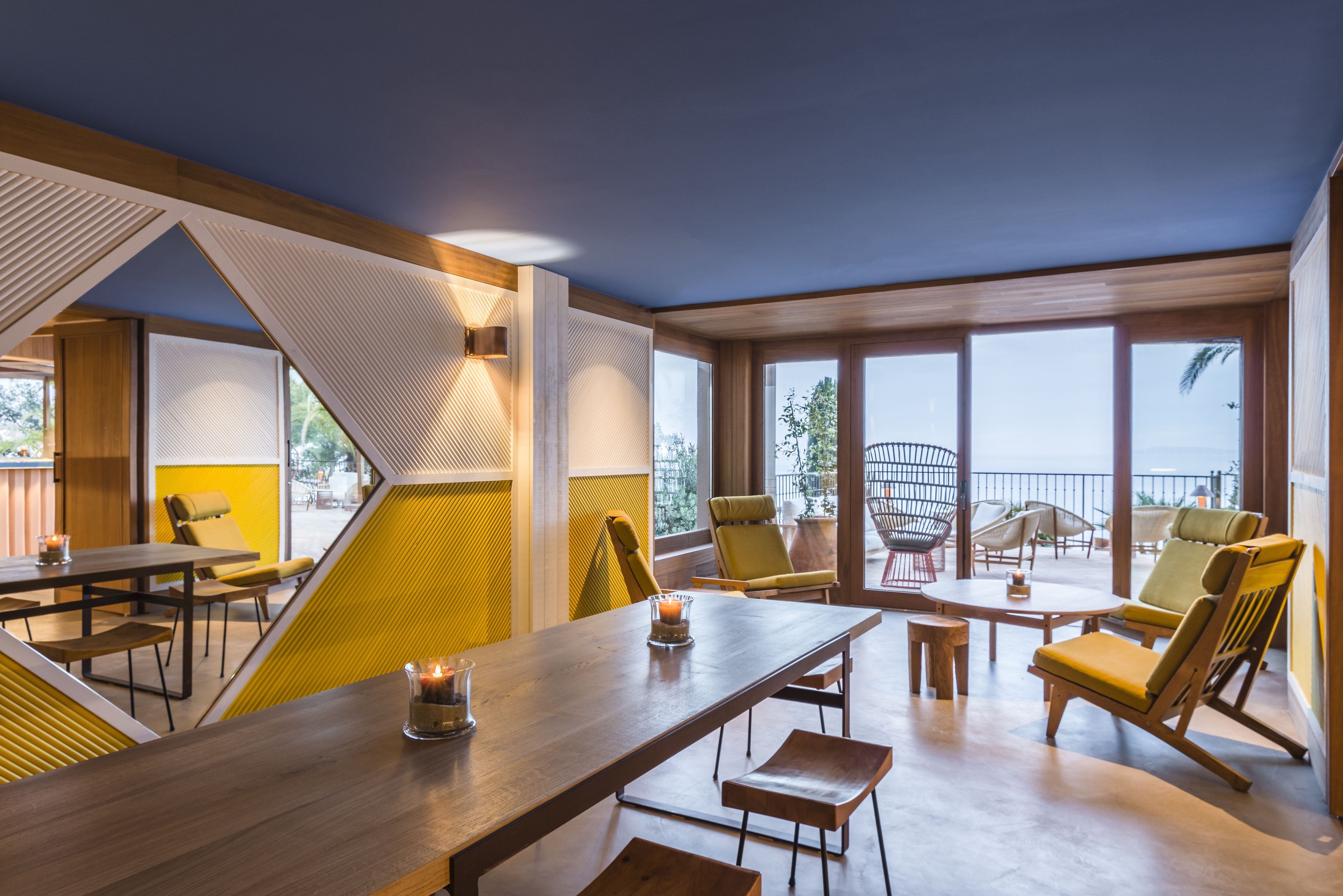 A modern dining area with a long wooden table, yellow chairs, and a view of the outdoors through large windows.