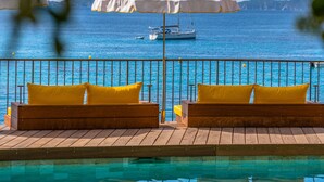 Zona junto a la piscina con tumbonas de madera y una sombrilla blanca, con vistas al mar azul y algunas embarcaciones.