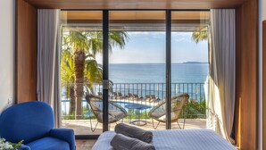 Habitación de hotel con un gran ventanal corredizo que da a un balcón con piscina y sillas, cama con toallas y vista al mar.